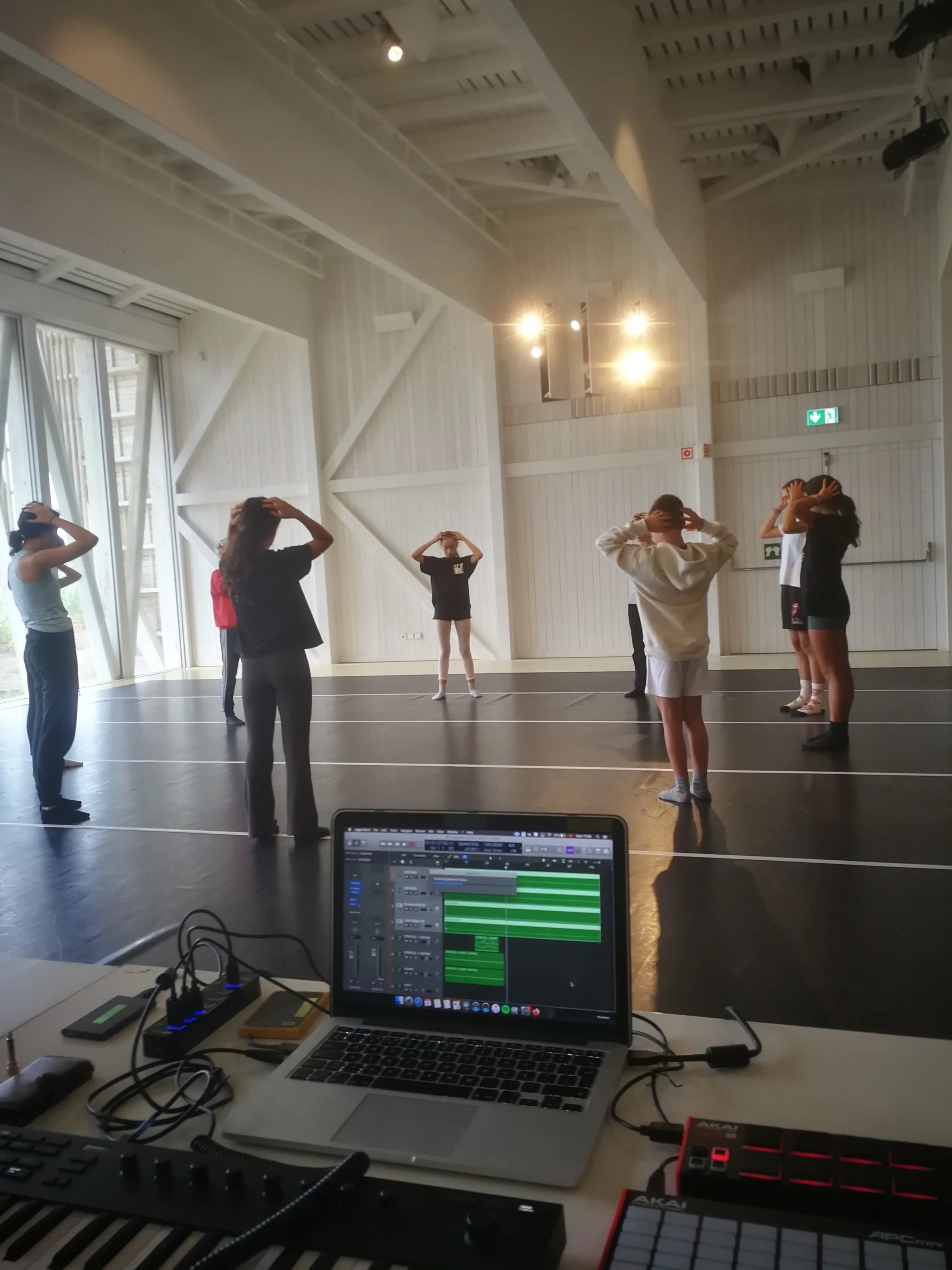 People in a dance studio practicing dance moves, with a person at the front leading, while a person in the middle acts as the instructor. The studio has high ceilings, large windows, and bright lighting, with some dance equipment and a laptop with mu