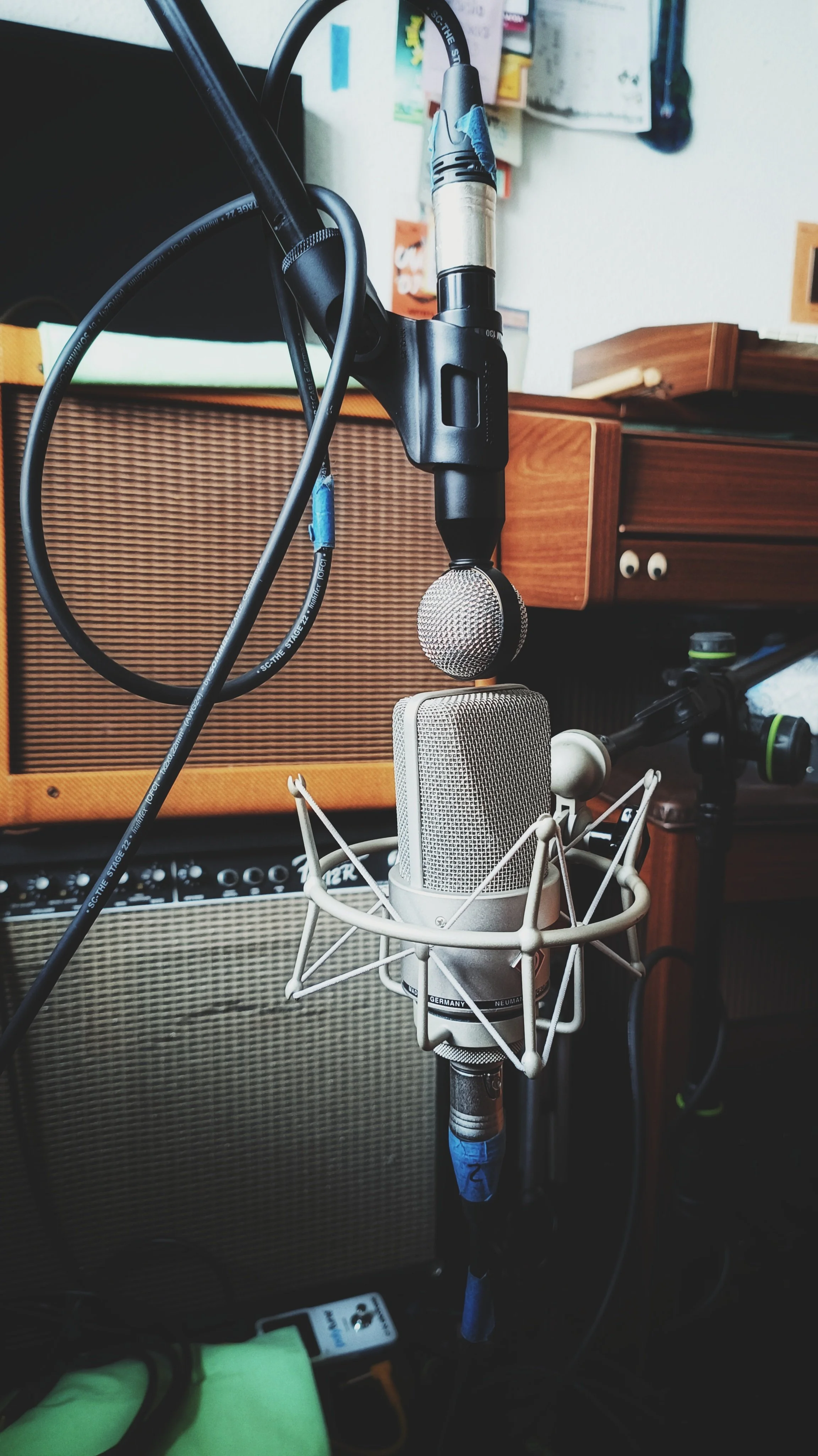A professional condenser microphone with a pop filter in a recording studio, with audio equipment and a guitar amplifier in the background.