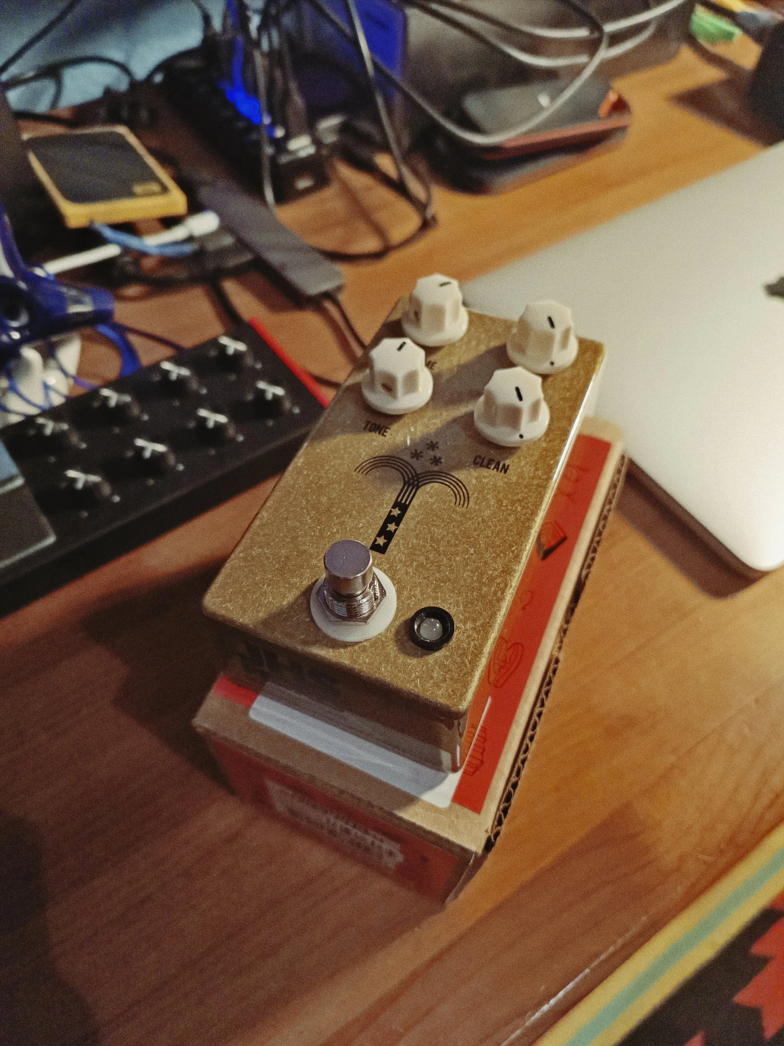 Close-up of a guitar effect pedal with four white knobs labeled 'Tone', 'Volume', 'Gain', and 'Clean' on a wooden surface, with various electronic devices and wires in the background.