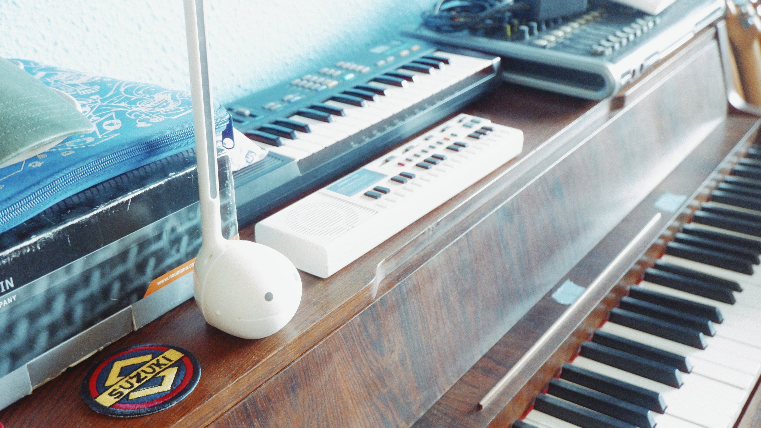 A collection of musical instruments on a wooden surface, including a keyboard, a small synth, and a laptop with a mixing console, with a patterned mug and a small bird-shaped object nearby.