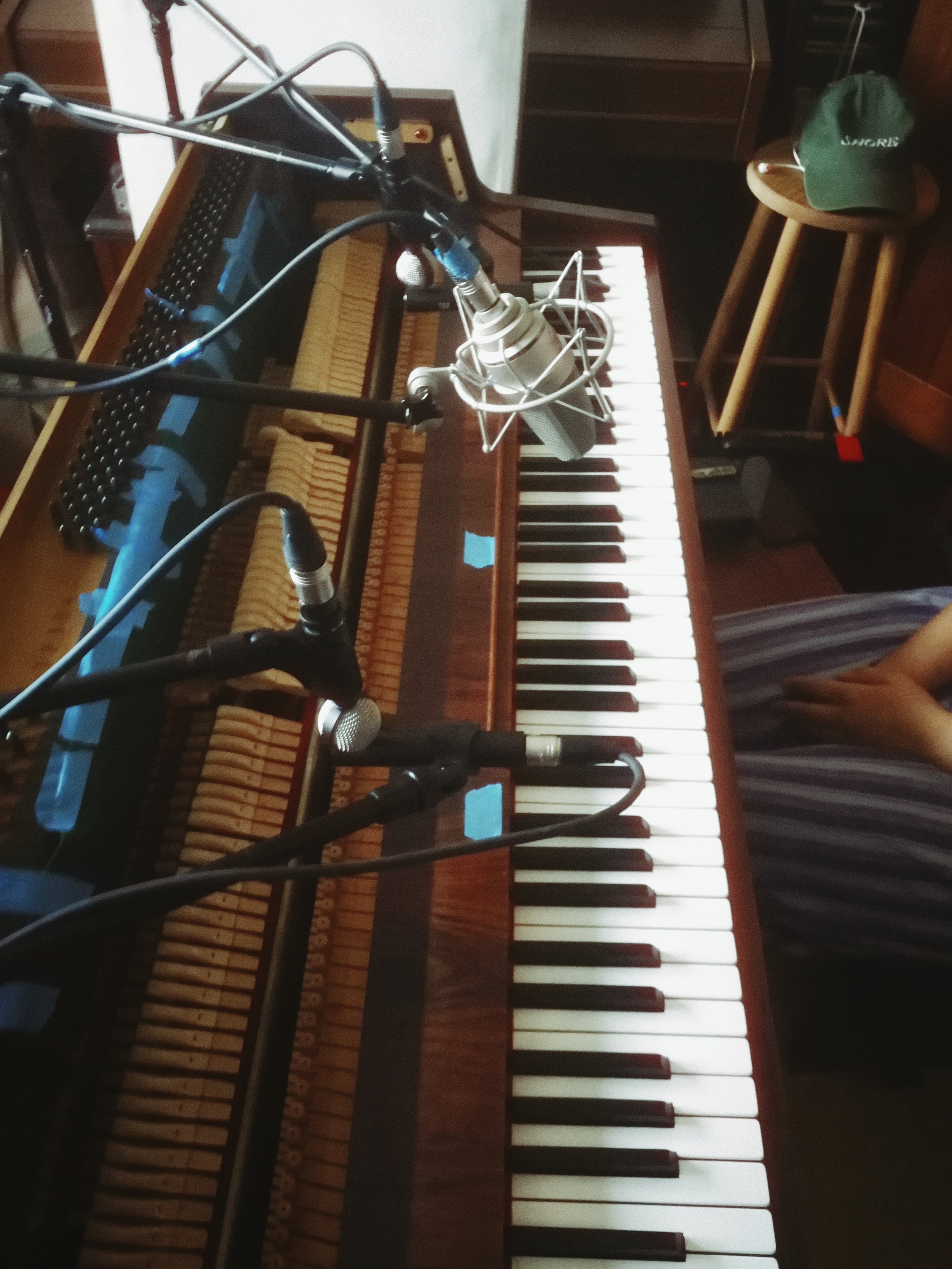A recording studio setup with a vintage wooden electronic keyboard, a microphone on a stand, and a pair of headphones resting on a stool. There are some cables connected to the microphone and the keyboard.