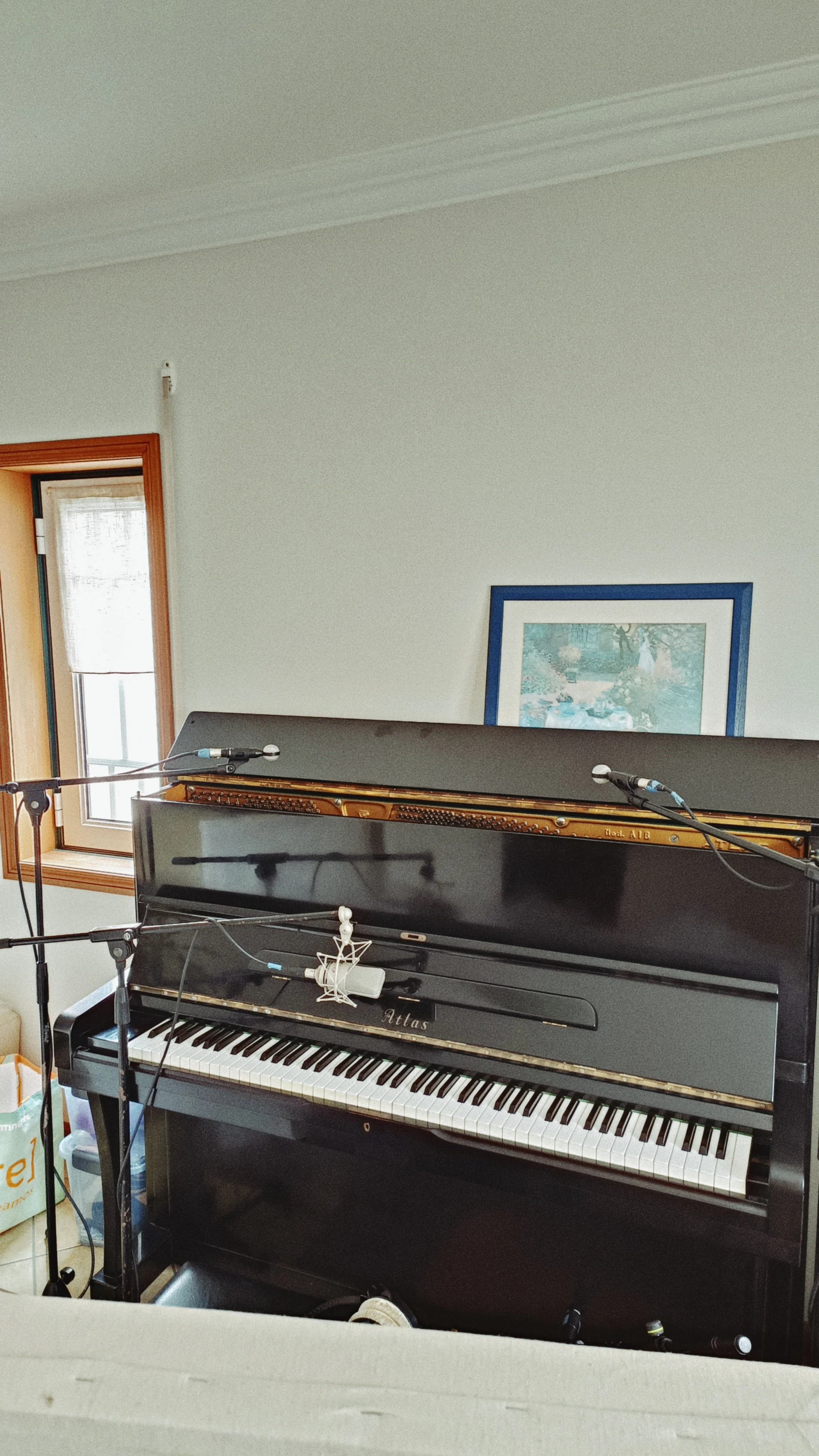 A black upright piano with a brown interior, microphones, and music equipment in front of it. A framed painting or photograph hangs on the wall behind, near a window with a wooden frame.