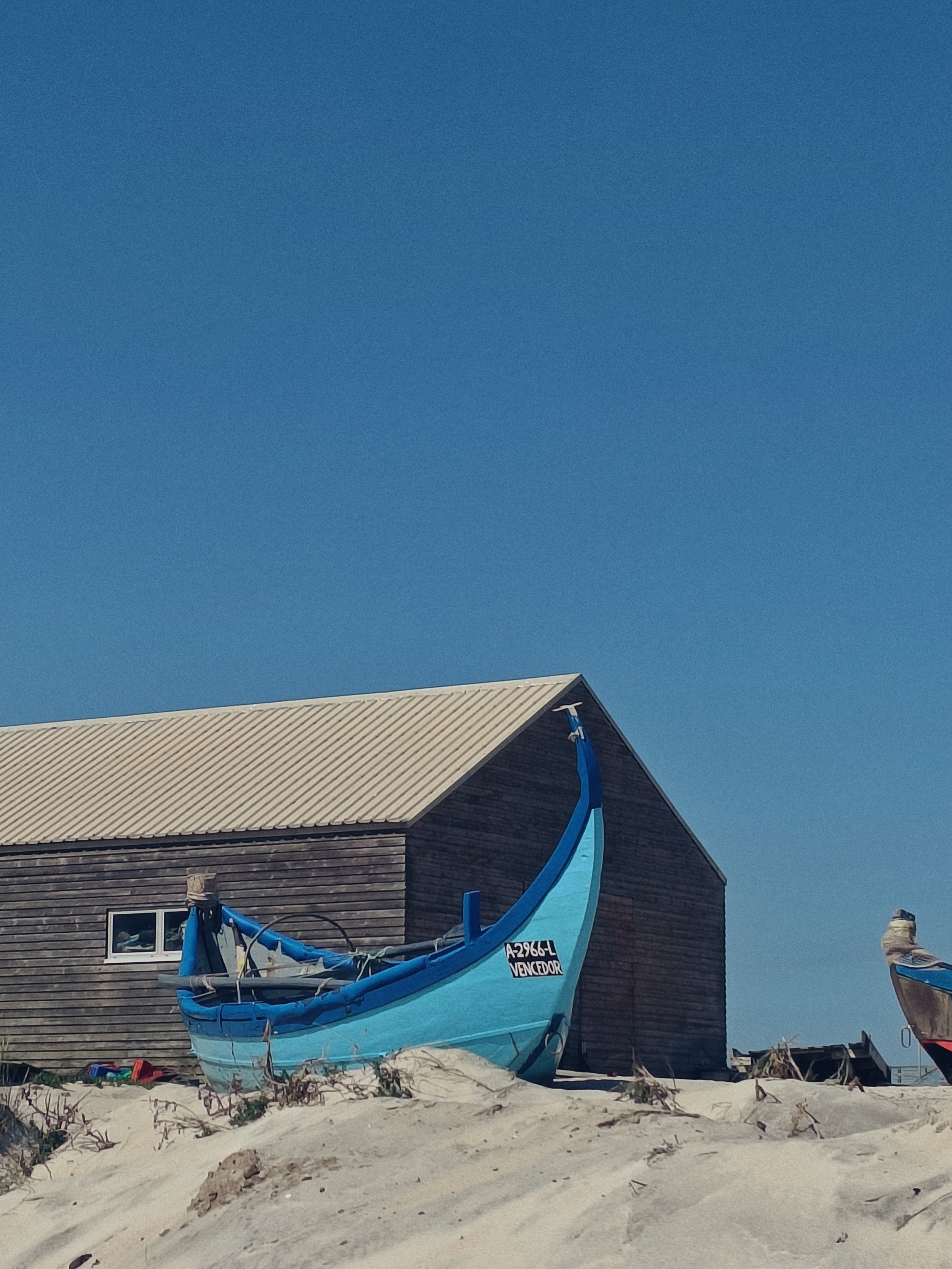 A small blue boat without a sail is grounded on sandy terrain in front of a wooden beach house with a beige roof, and the sky is clear and blue.