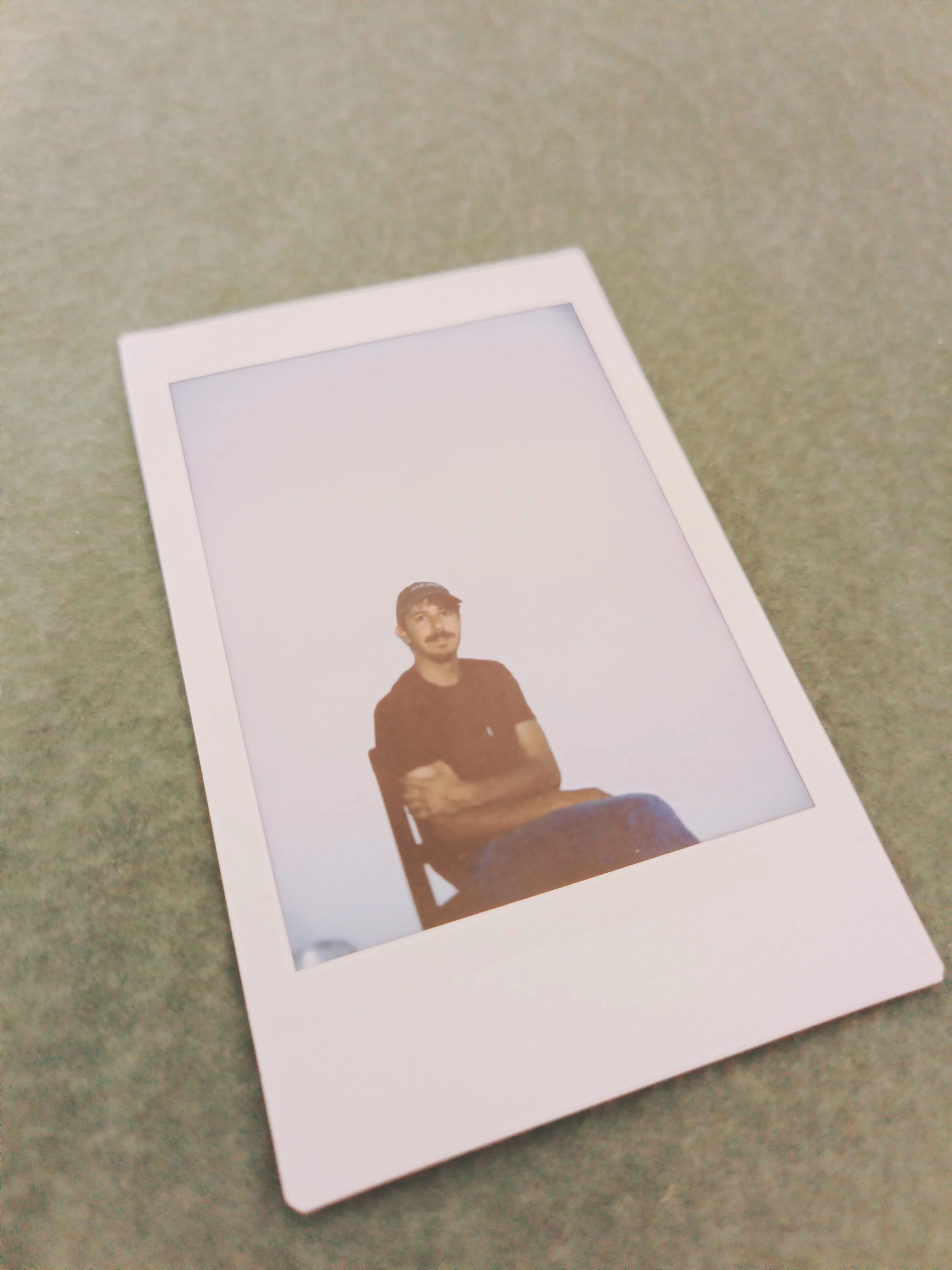 A photograph of a Polaroid photo featuring a man with a mustache, wearing a dark shirt, sitting on a chair with his hands resting on his lap, against a light background.