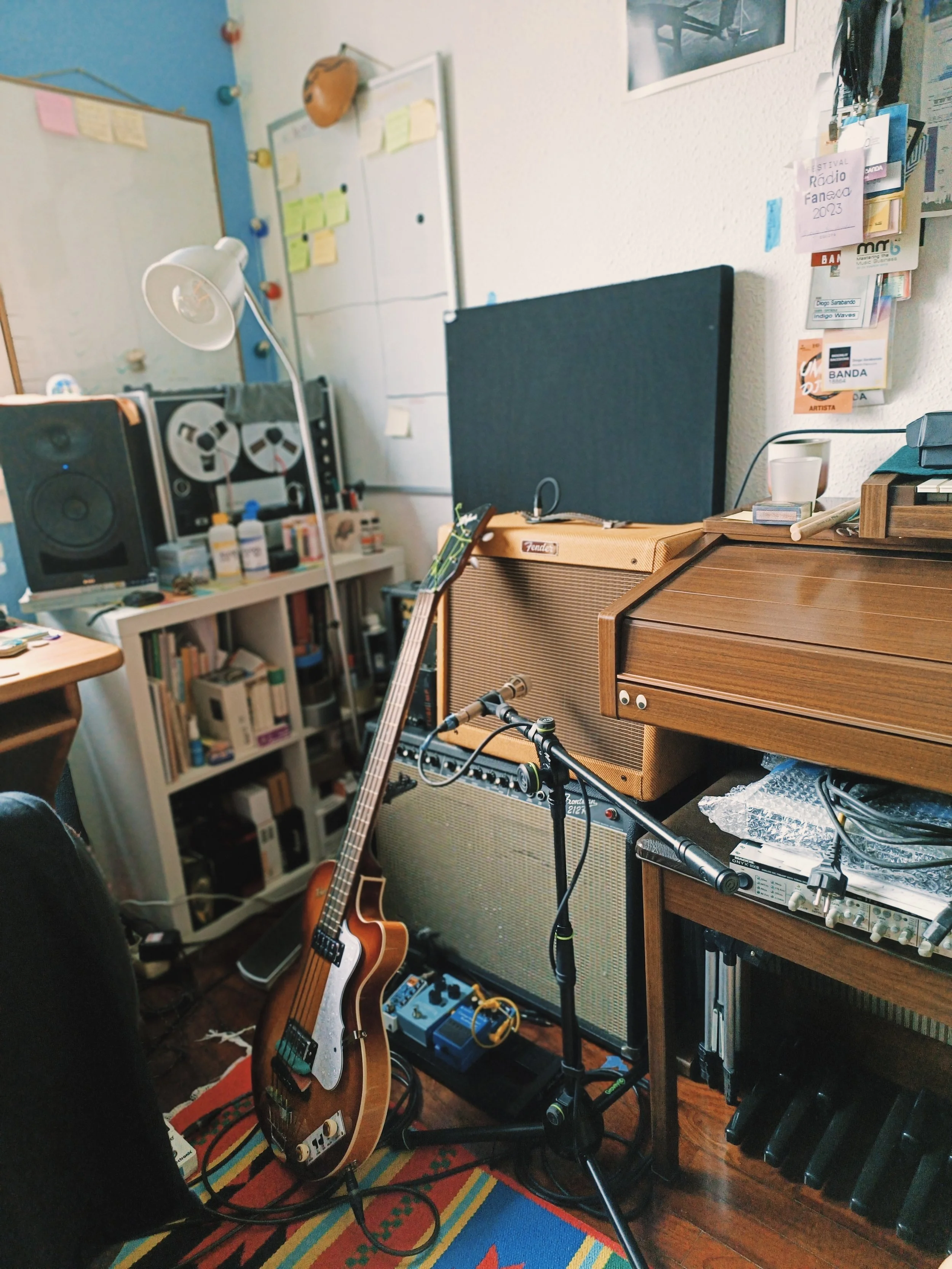 A room with musical equipment, including a guitar, an amplifier, a desk with a speaker, and cds. There are shelves with books and a wall decorated with Post-it notes and a poster.