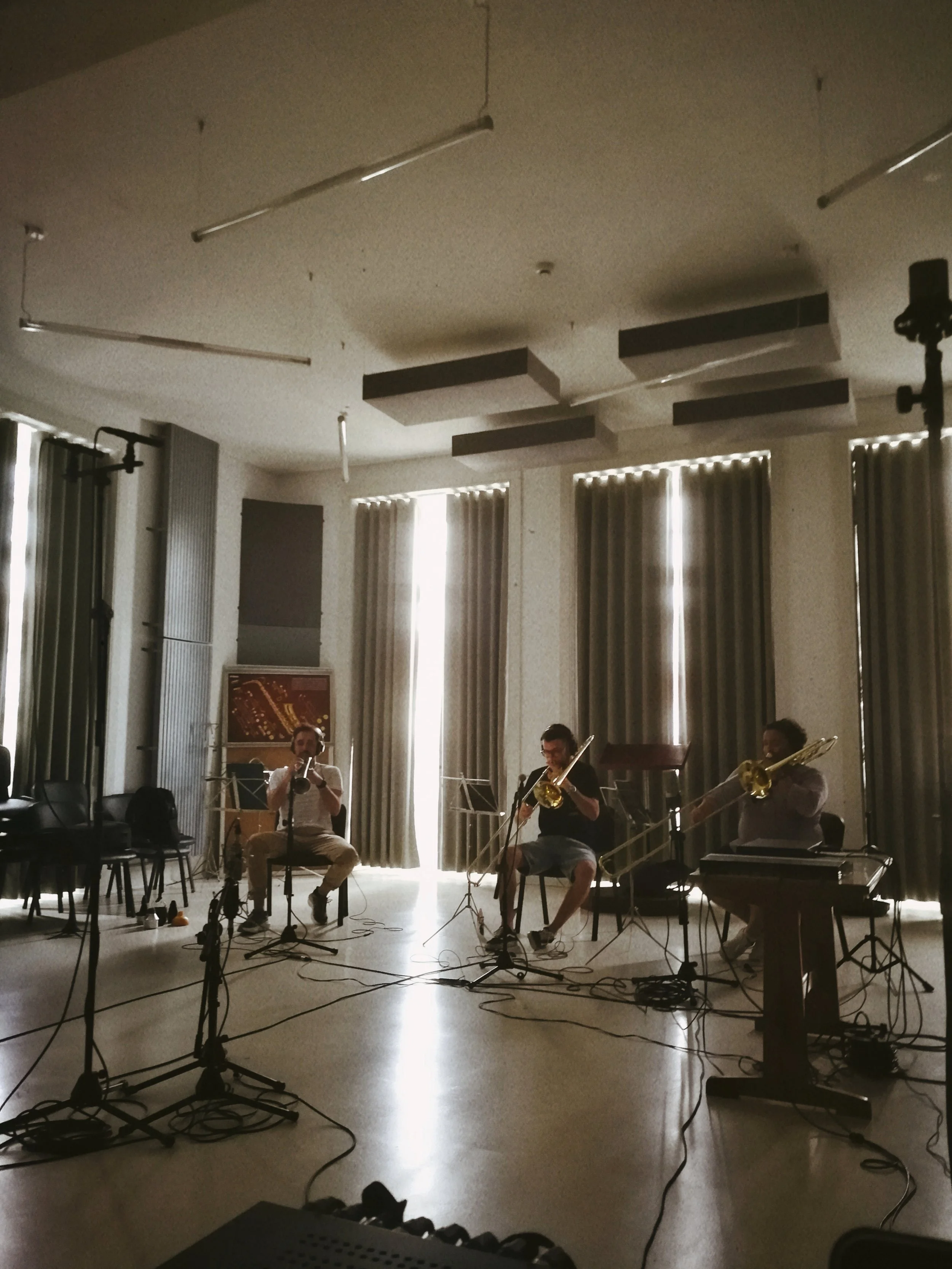 Musicians playing instruments in a dimly lit room with large windows and curtains, surrounded by microphones and equipment.