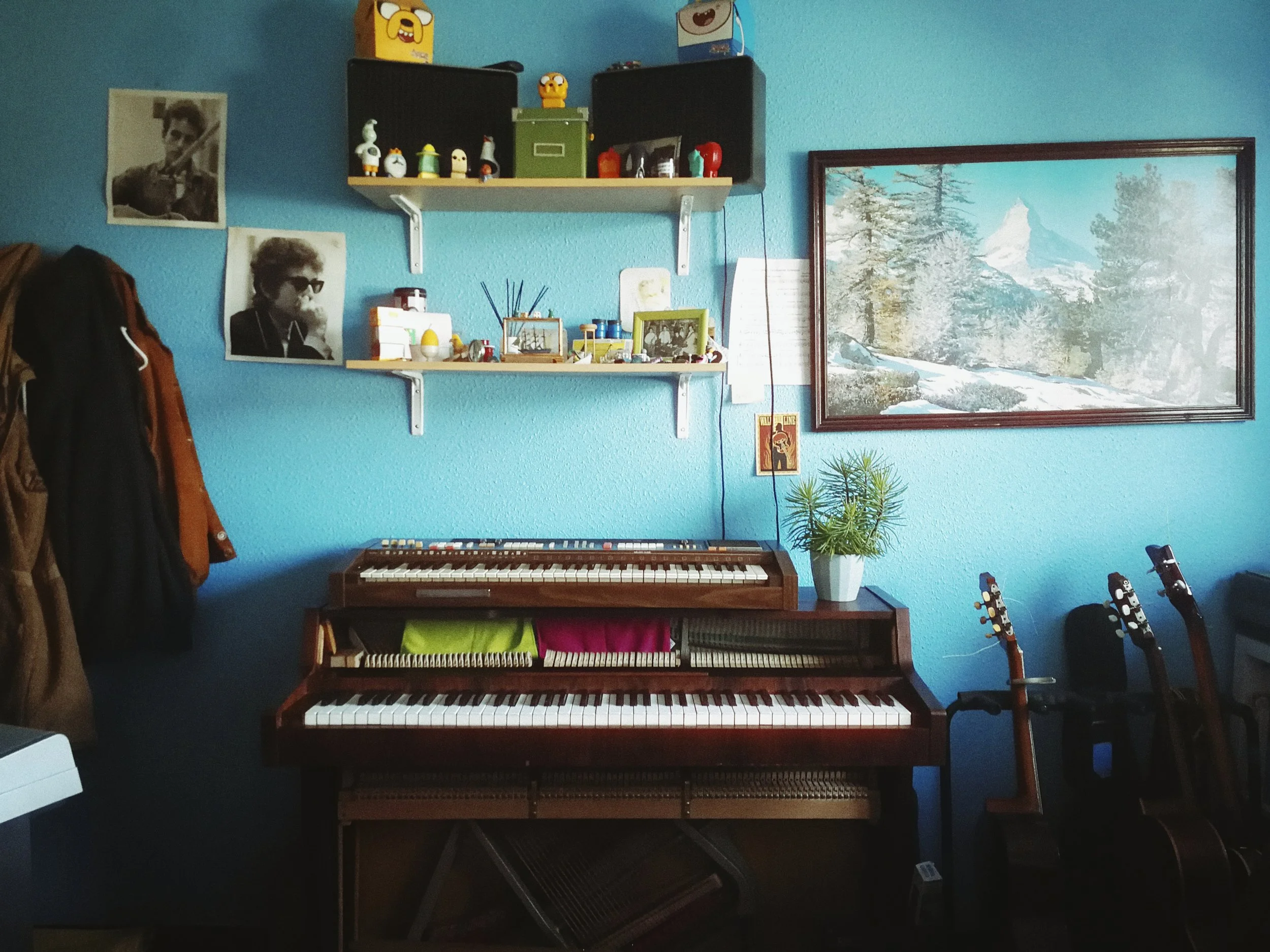 A room with a blue wall featuring two black shelves filled with small figurines, some framed pictures, and decorative items. There is a large framed landscape painting of snowy mountains and pine trees on the wall. Below the shelves, a digital keyboa