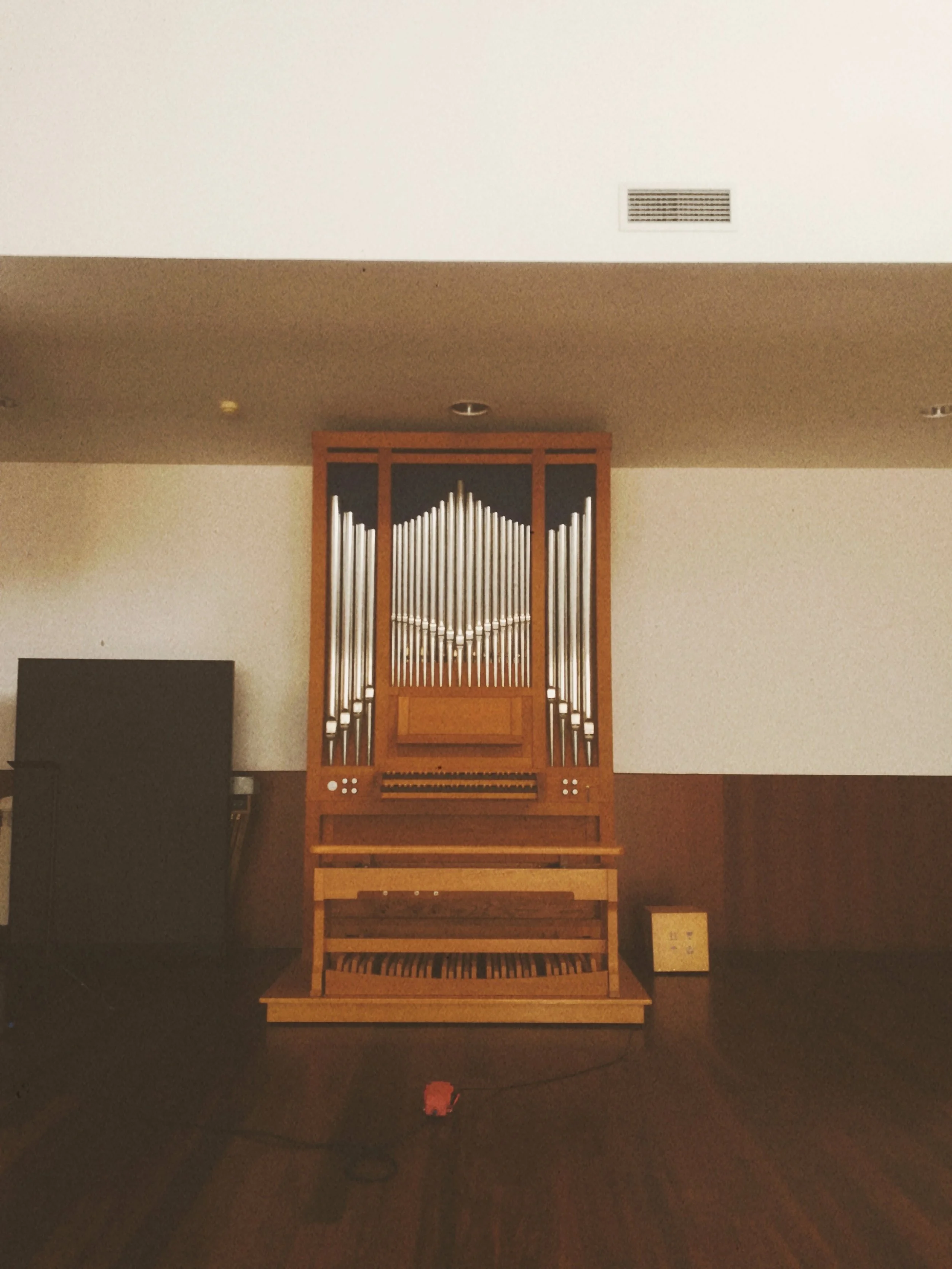 A large pipe organ with metal pipes mounted on a wooden frame, situated against a beige wall in a dimly lit interior space.