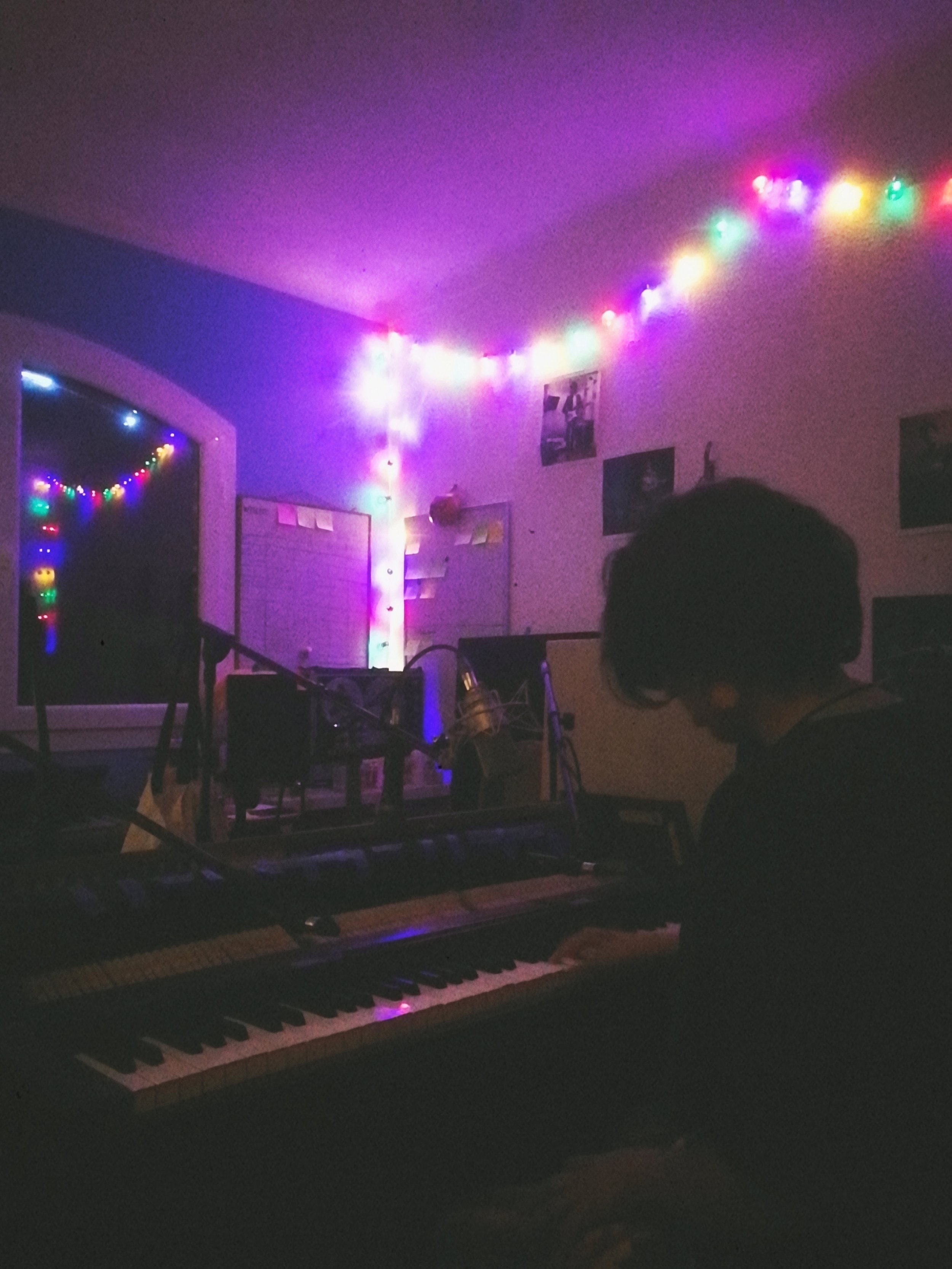 Room decorated with multicolored string lights, a piano with a keyboard, and posters on the wall, with a person playing the piano surrounded by musical equipment.