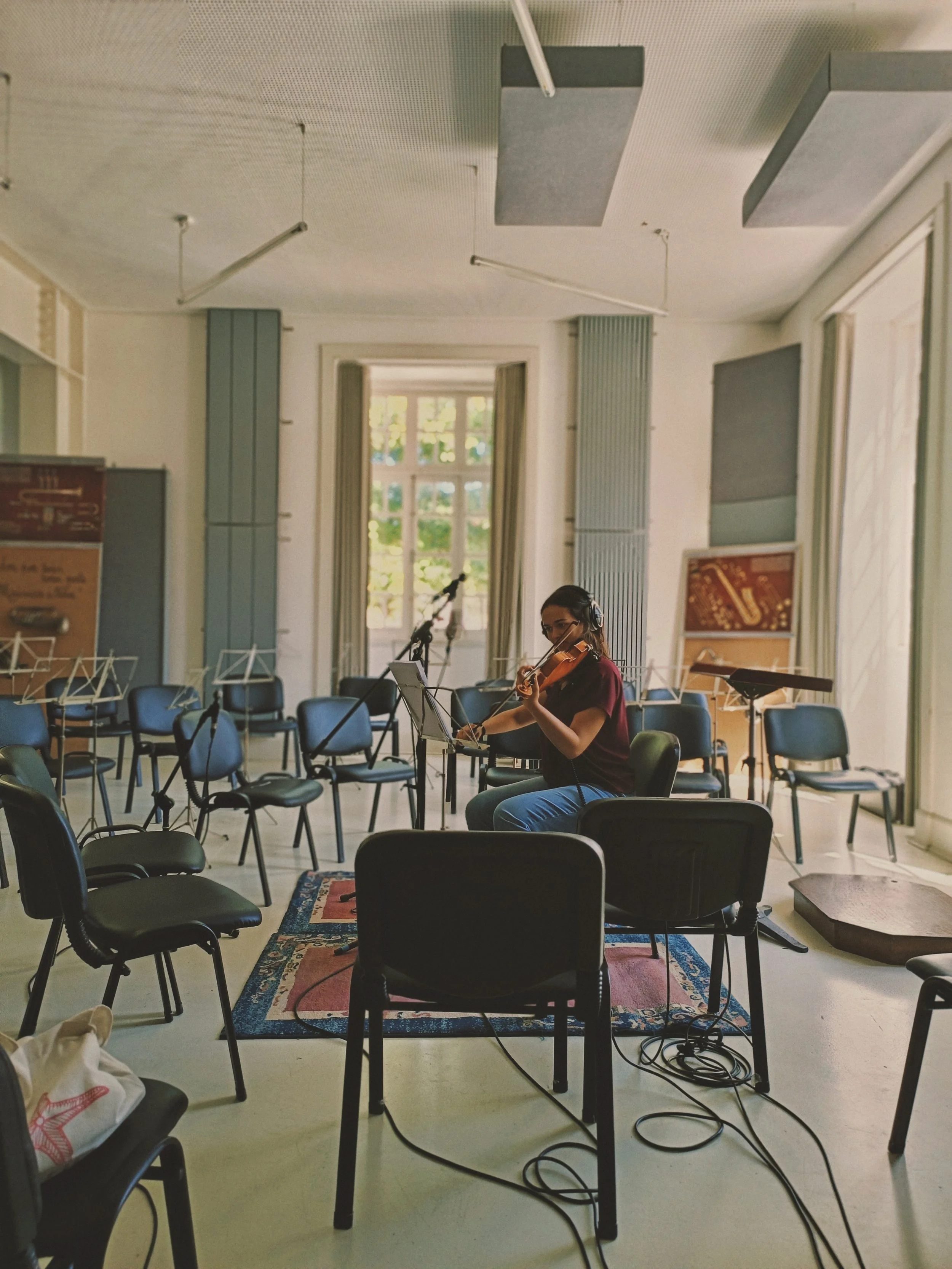 A woman playing the violin in a room with chairs arranged for a performance, windows letting in natural light, and music stands present.
