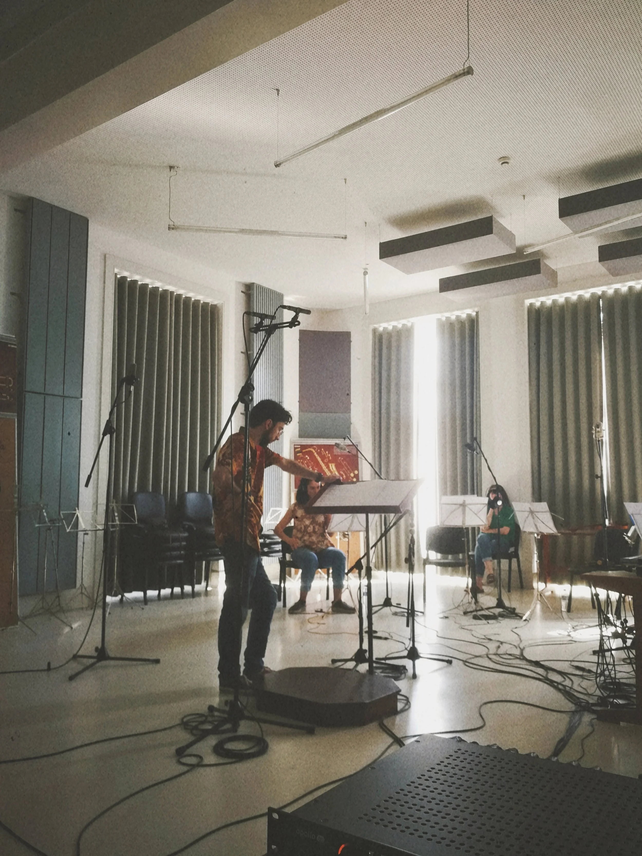 A group of people rehearsing or performing on a stage with music stands, microphones, and chairs, in a room with large windows and curtains.