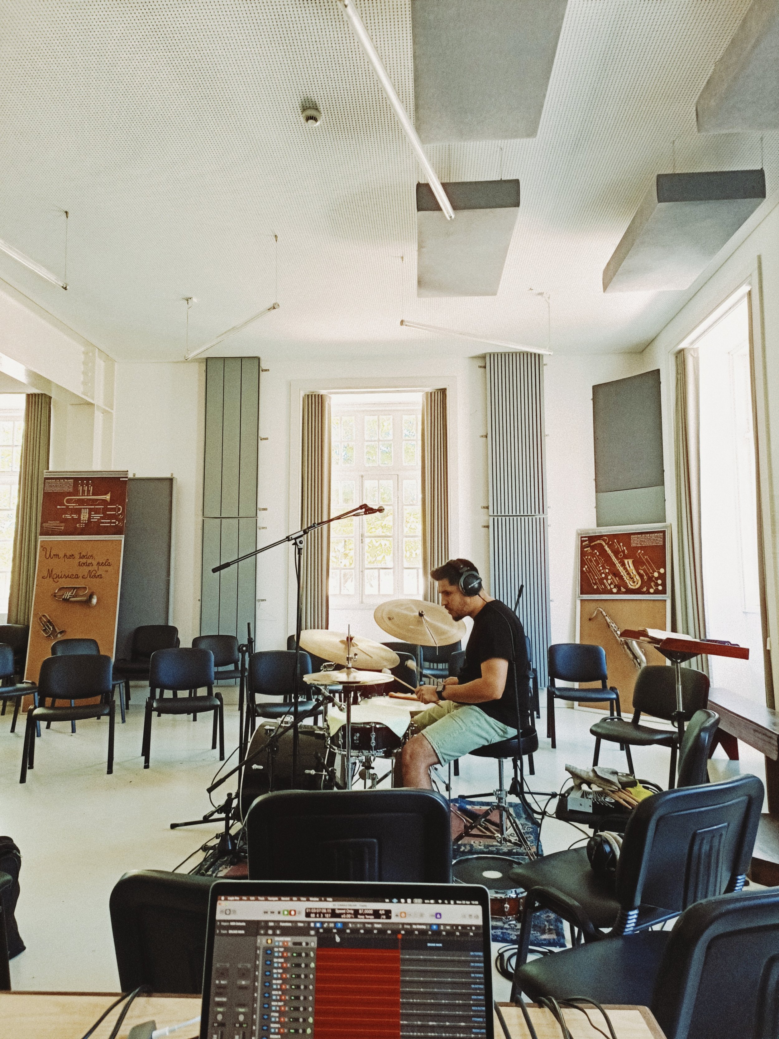 A man sitting on a chair playing drums, wearing headphones in a music rehearsal room with chairs, music stands, and musical equipment, large windows with curtains in the background.
