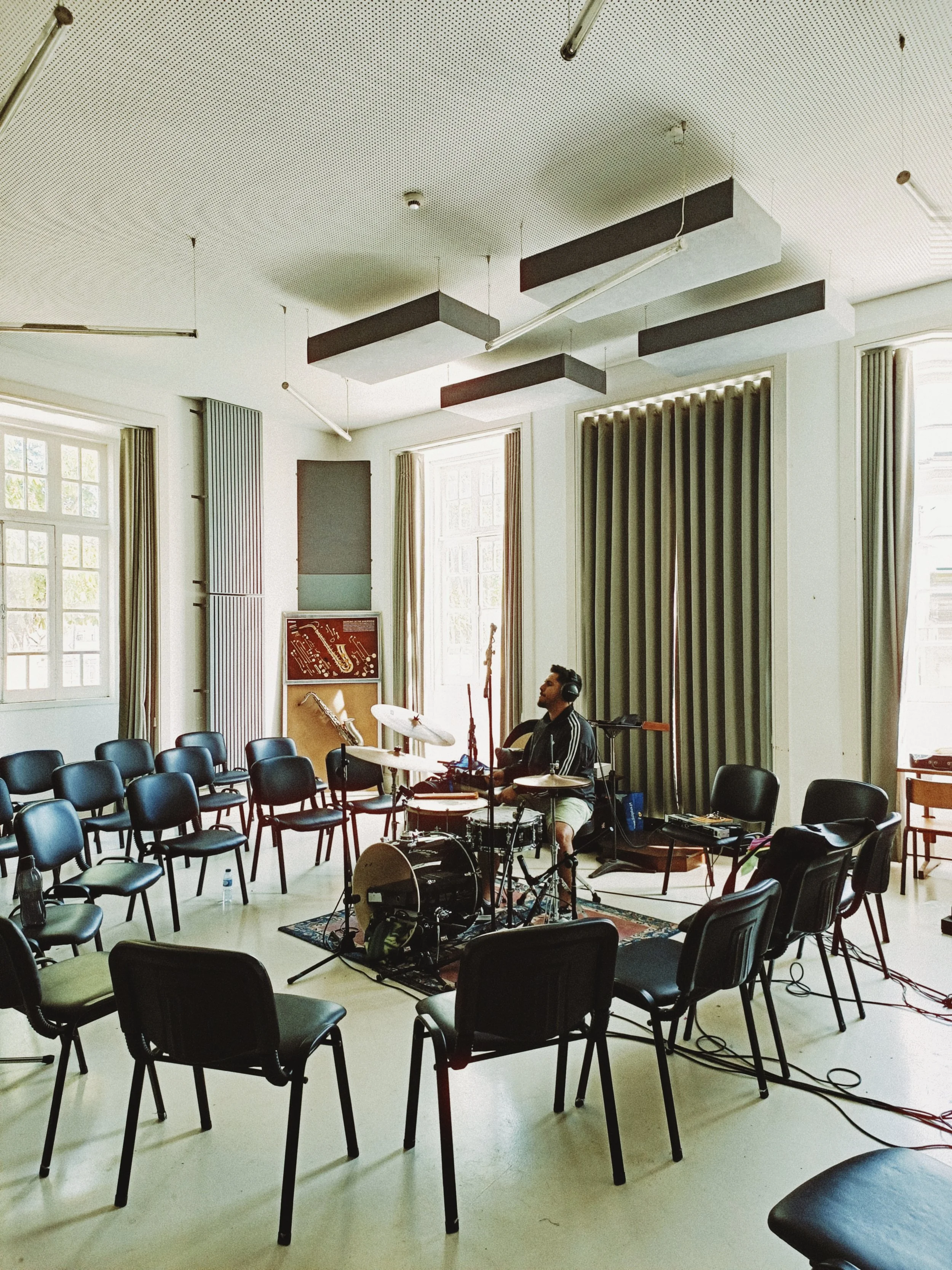 A man wearing headphones playing drums in a room with chairs arranged in a semi-circle, windows with curtains, and soundproofing panels on the walls and ceiling.