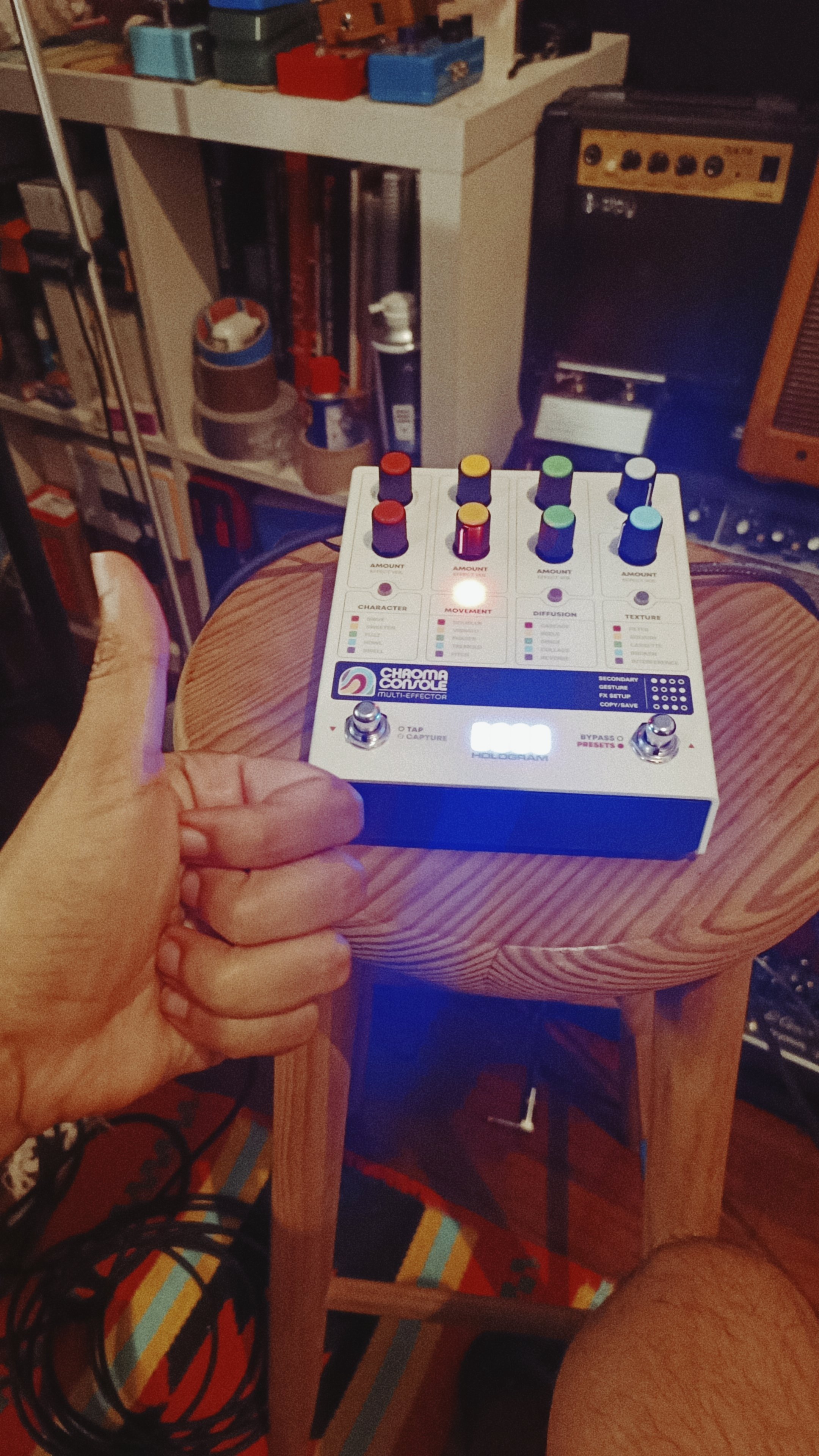A person holding a colorful electronic music device called a Chaomea Controller Multi-Effect, with various knobs and buttons, set on a wooden chair in a room with shelves and electronic equipment in the background.