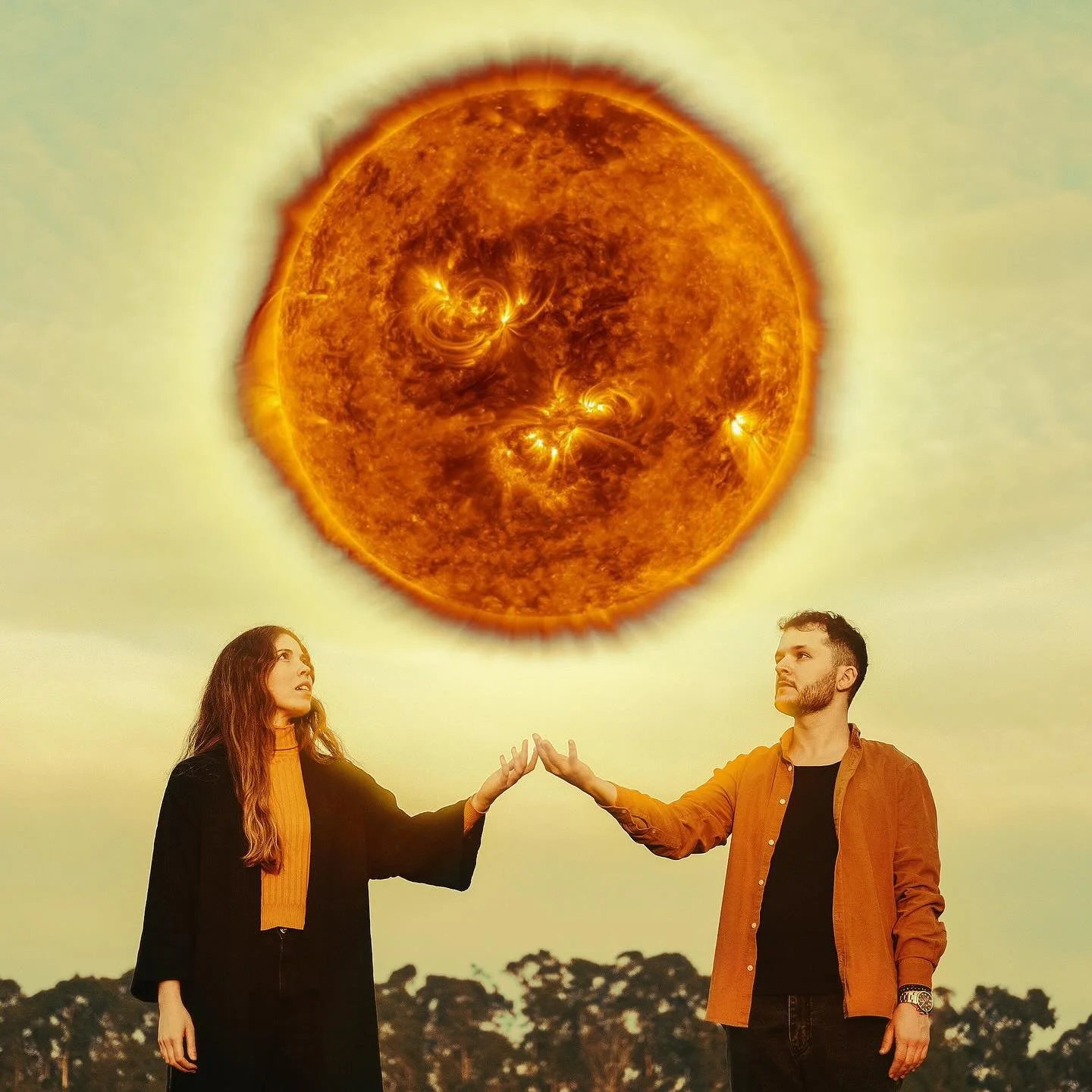 A woman and a man are standing outdoors with trees in the background, reaching towards a large, digitally manipulated sun in the sky.