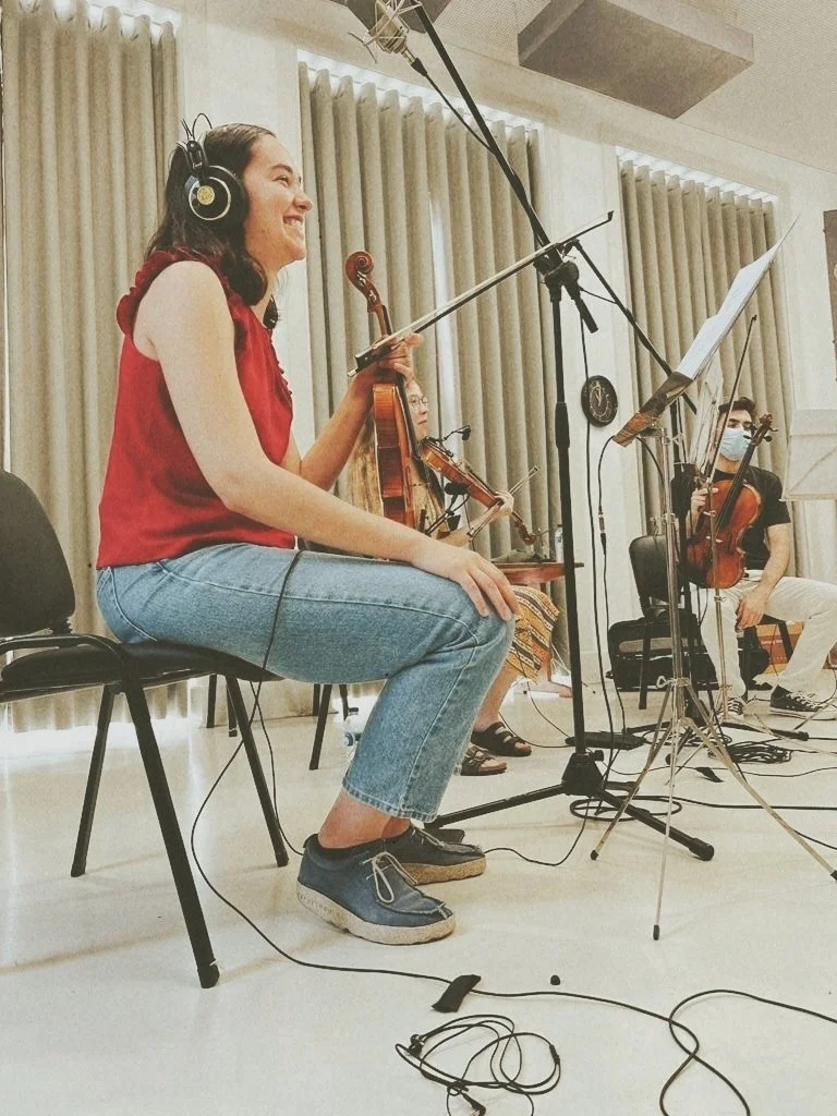 A woman with long dark hair, wearing a red sleeveless top, blue jeans, and gray shoes, is sitting on a chair playing a violin in a recording studio. She is smiling and wearing headphones. Other musicians with violins are visible in the background. Th