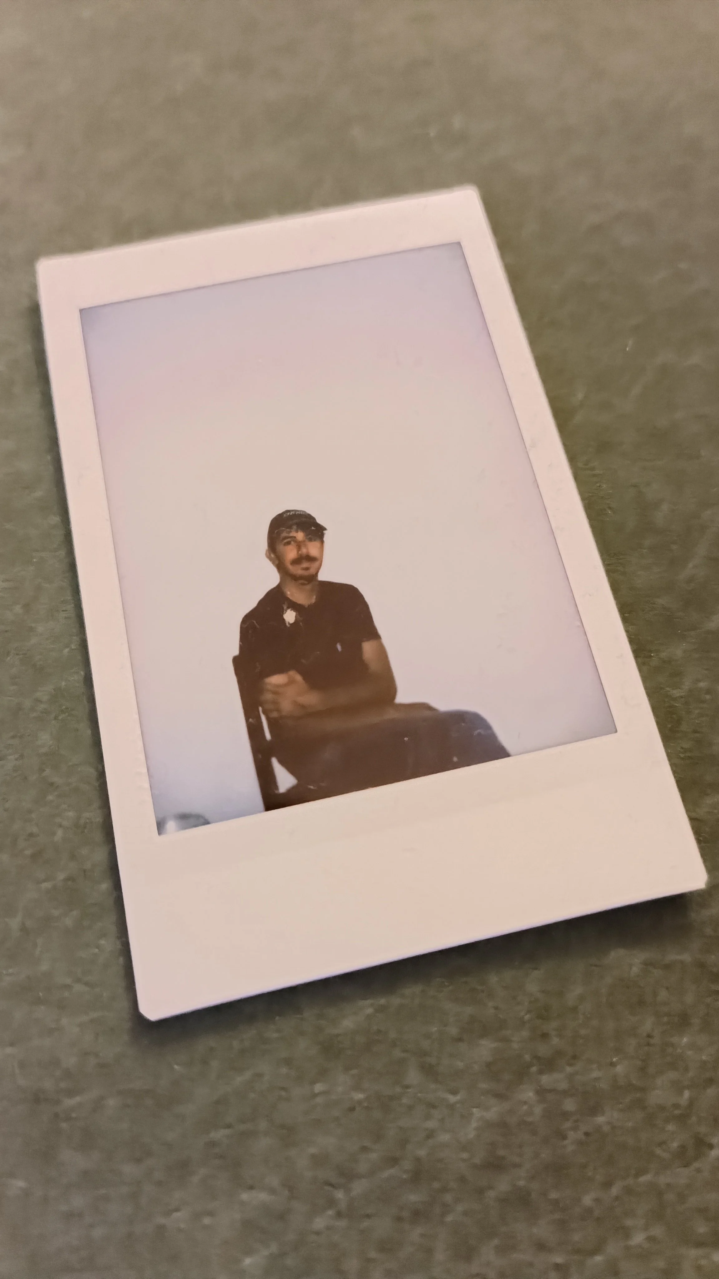 A Polaroid photo of a man sitting on a chair with his arms crossed, wearing a dark shirt, against a plain light background.