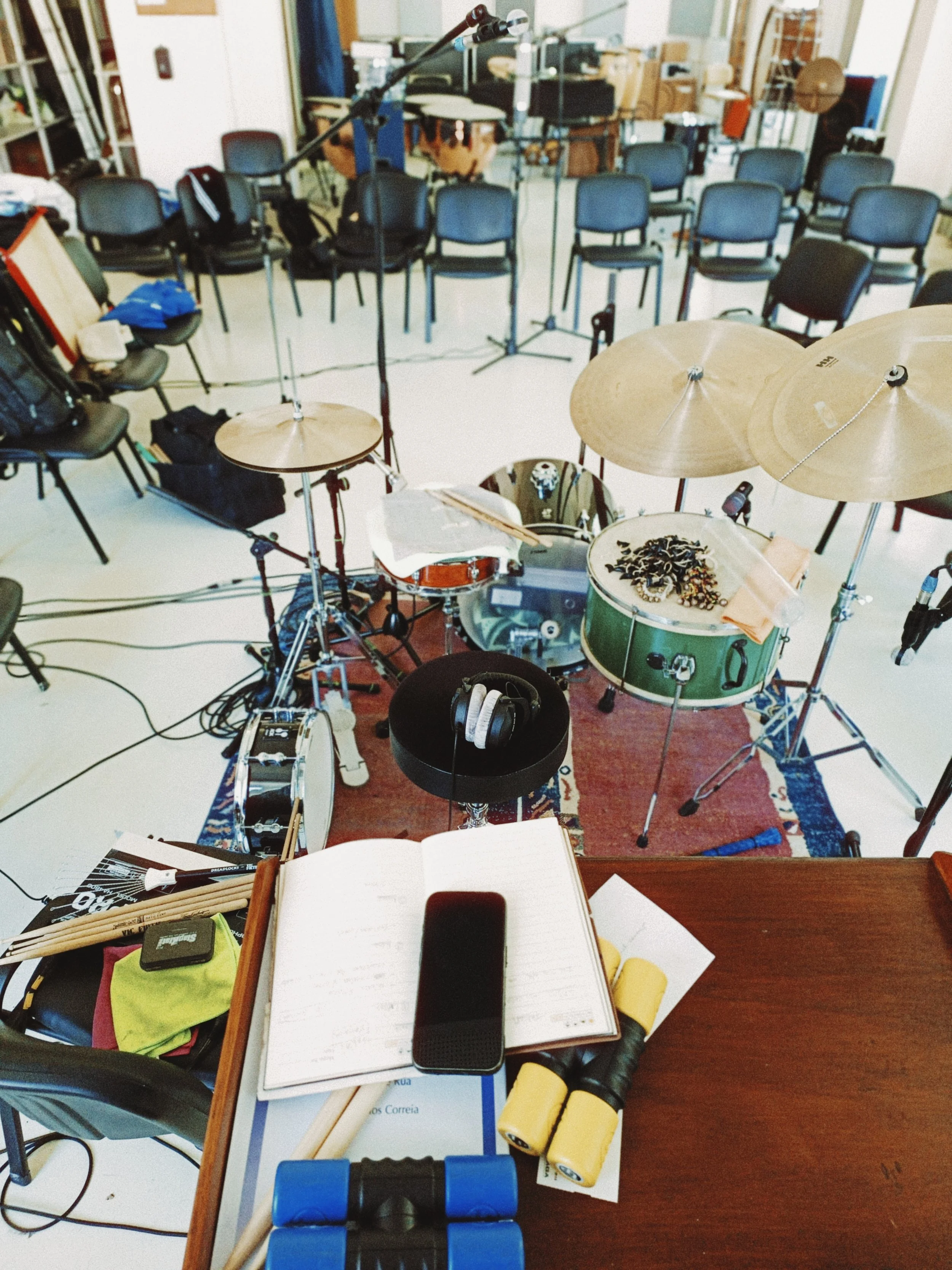 View from a drum kit facing towards empty black chairs arranged in rows, with music stands and equipment in the background, and a table with music sheets, headphones, and yellow batteries in the foreground.