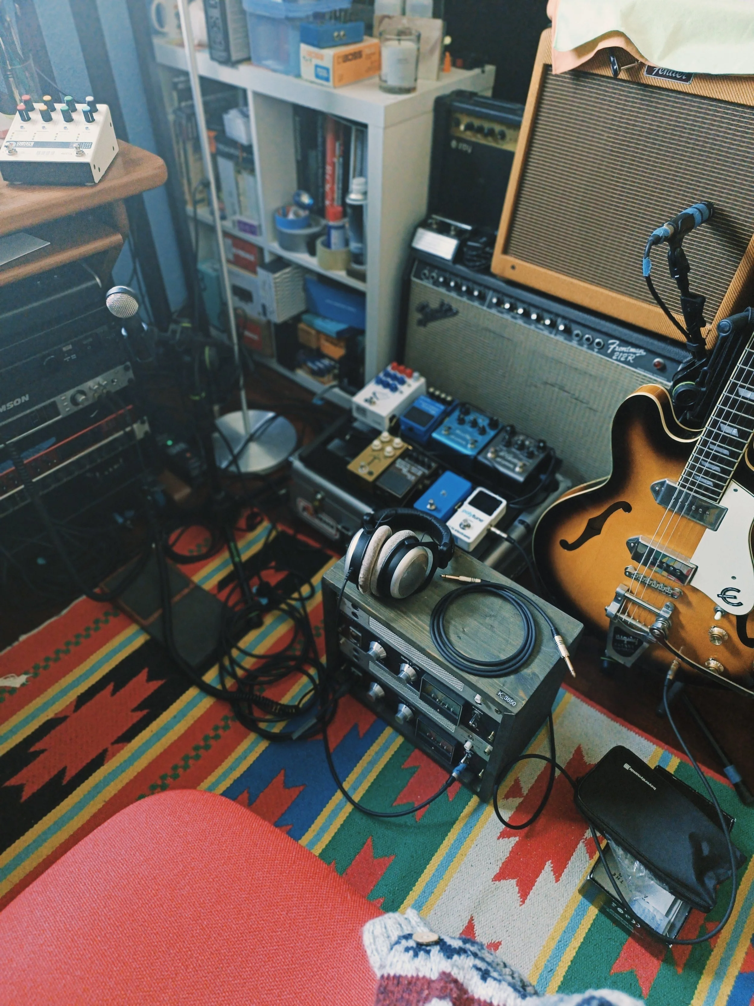 A recording studio setup with musical equipment including a guitar, guitar amplifier, microphone, and various audio gear, situated on a colorful patterned rug.