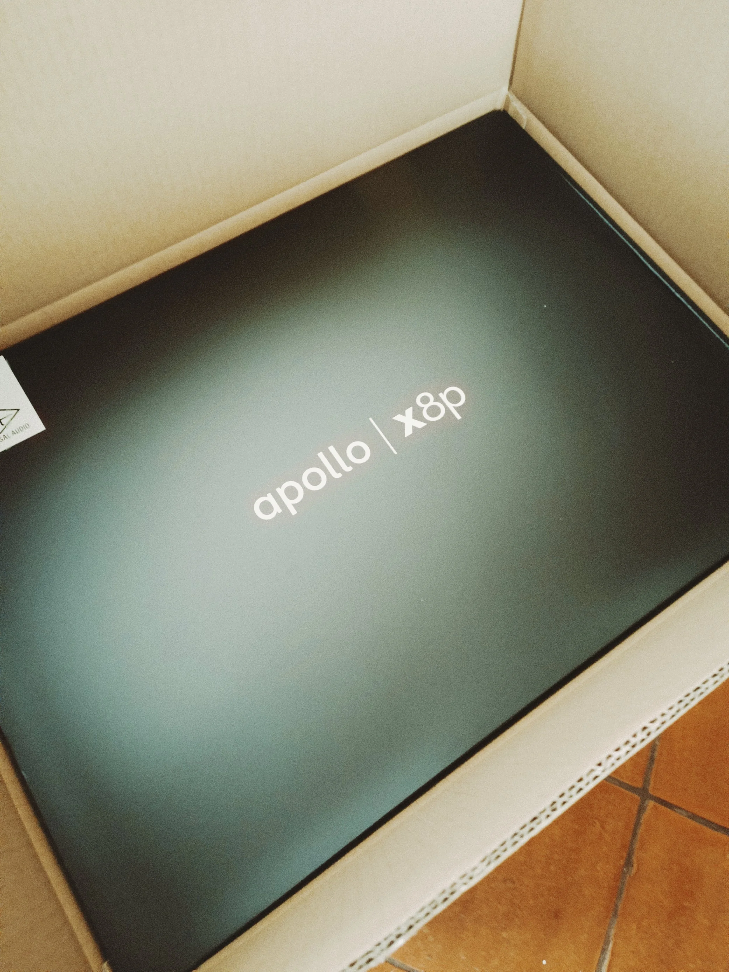 Close-up of a black kitchen appliance box with white text reading 'Apollo | x8p' on top, placed inside a cardboard box. Part of a white kitchen counter and brown tiled floor are visible.