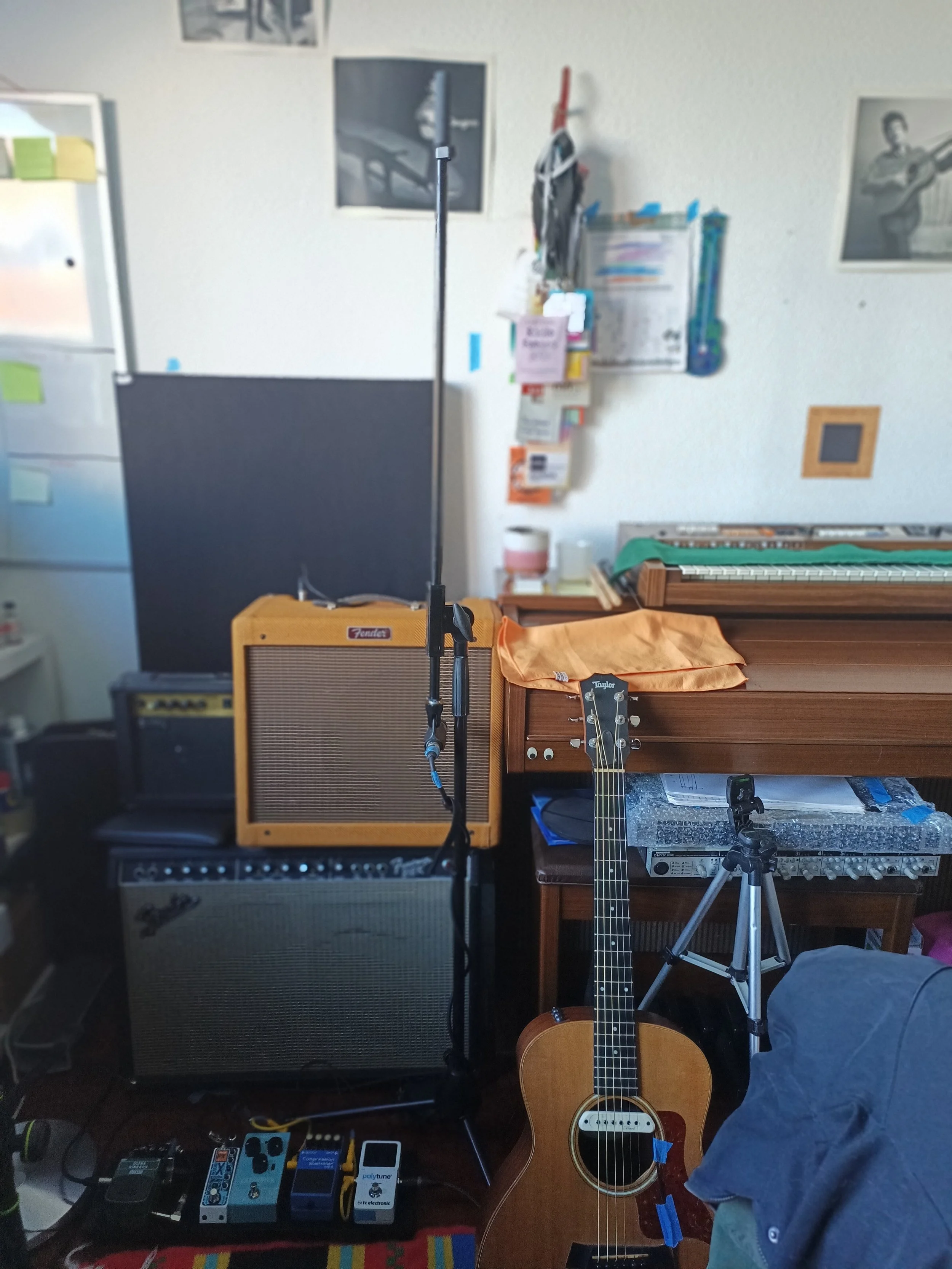 A living room corner with musical equipment including an acoustic guitar, a Fender amplifier, and several guitar effects pedals on a pedalboard, along with a microphone stand, a note on a wall organizer, and framed photos or artwork on the wall.