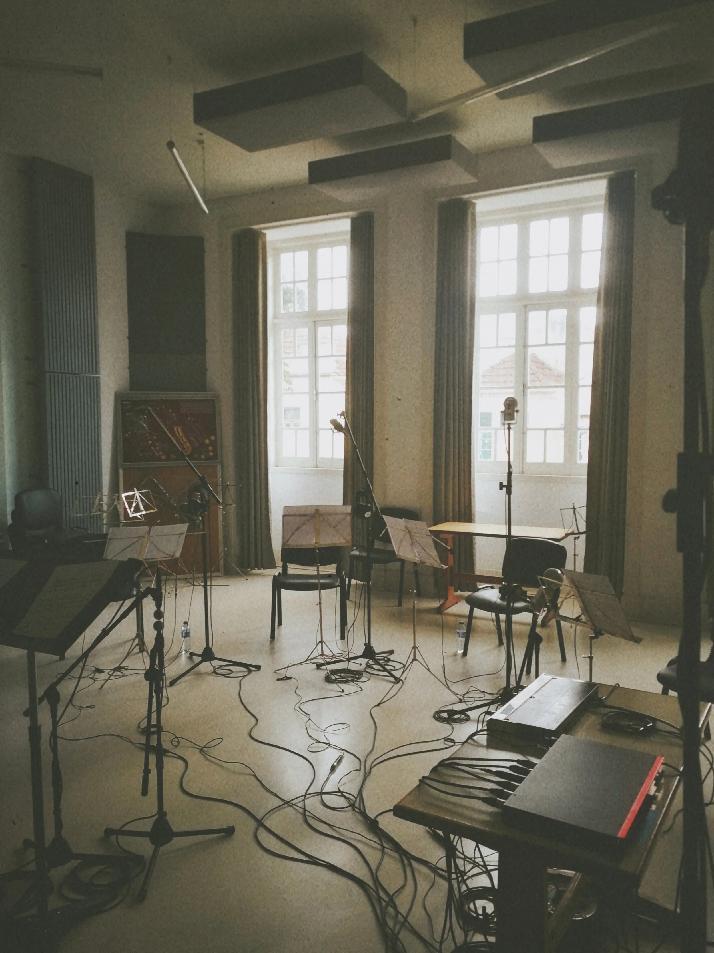 A music practice room with sheet music, microphones, a keyboard, and chairs arranged near large windows with curtains, with sunlight illuminating the room and a view of rooftops outside.