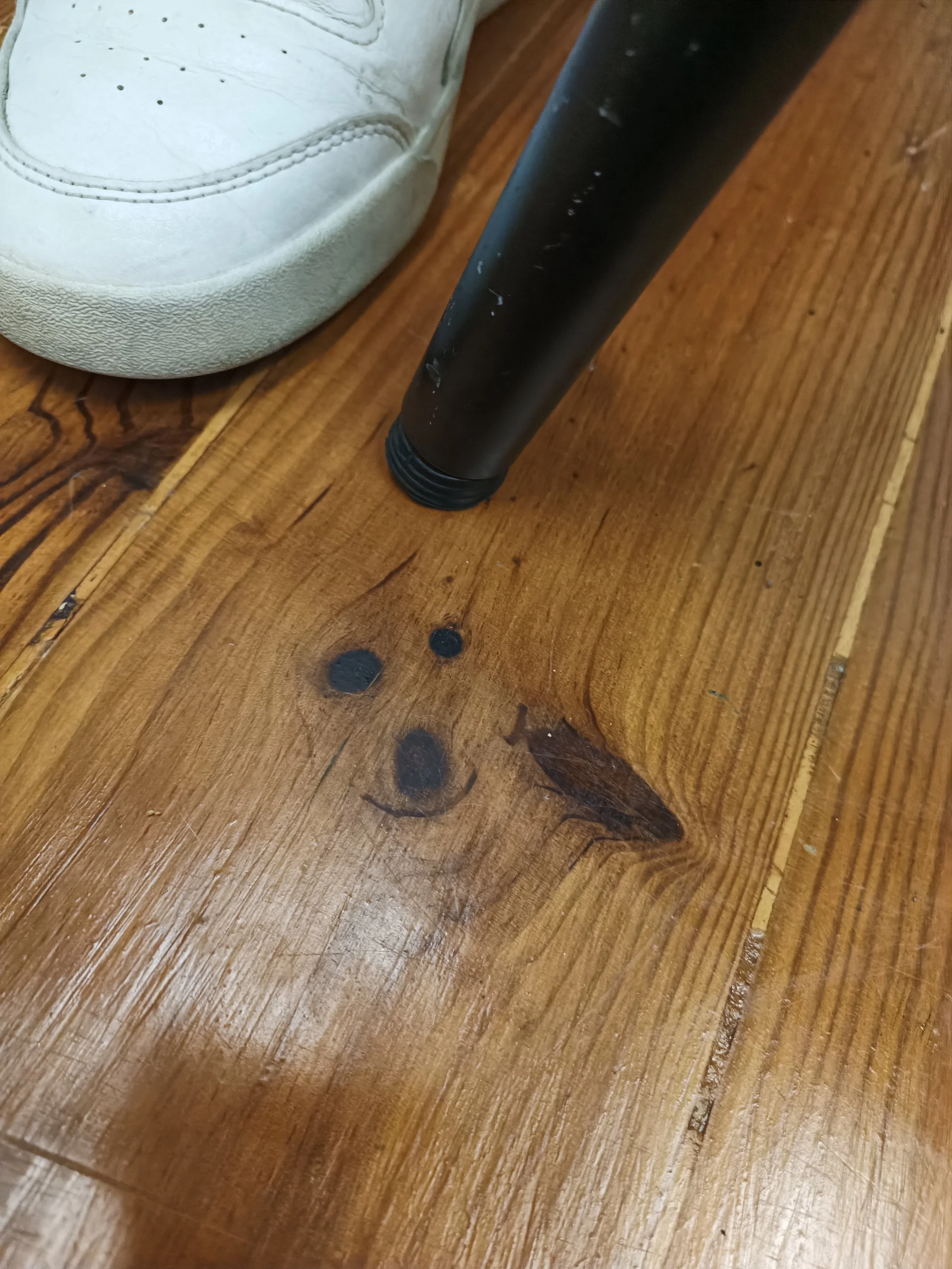 Close-up of a wooden table with a black metal leg, a white sneaker, and some black spots on the wood.