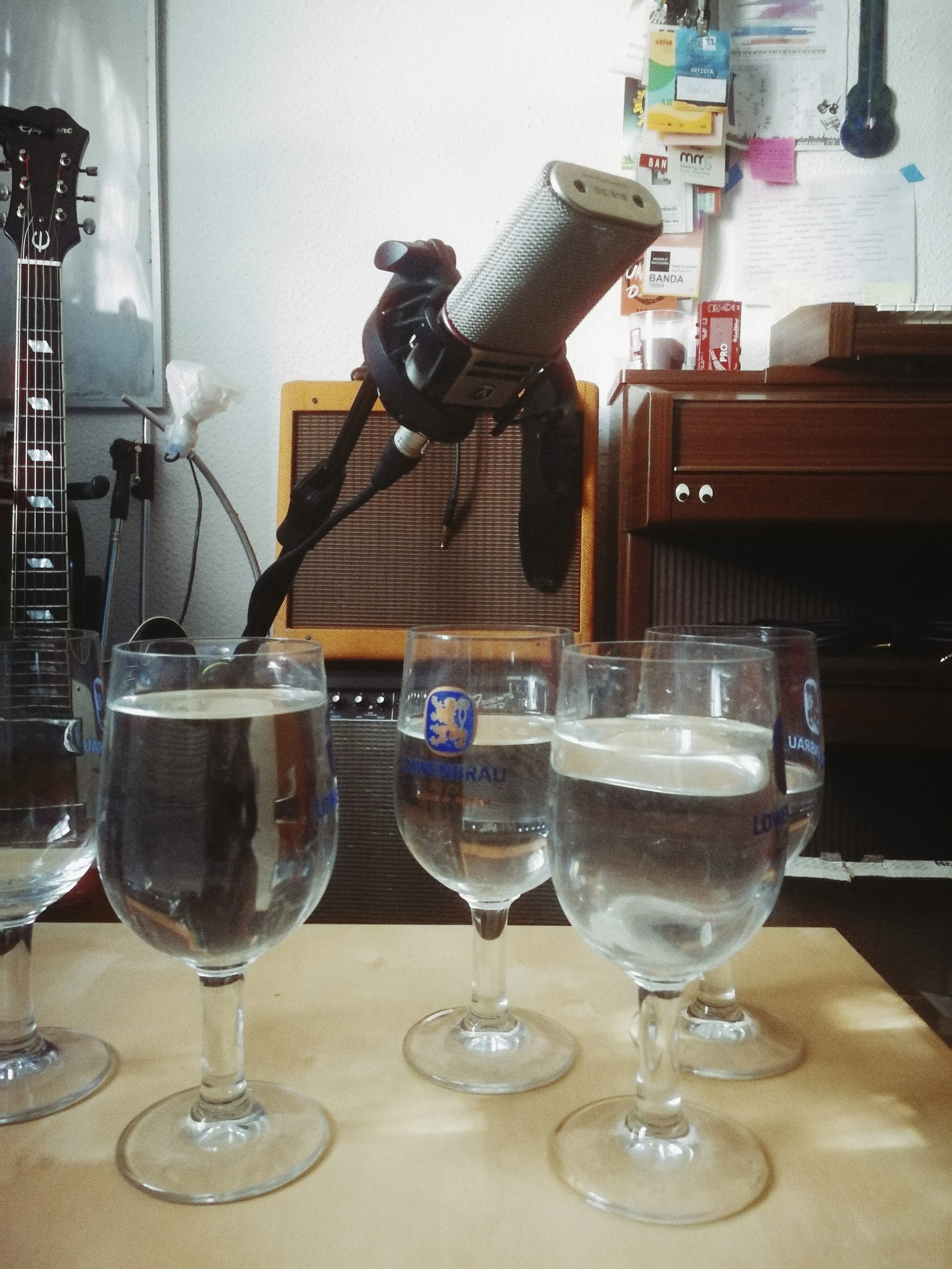 Six glasses of water or clear liquid placed on a table in front of a collection of variously colored glasses, bottles, or containers in a room with musical instruments, an amplifier, a speaker, and a wall with posters and notes.