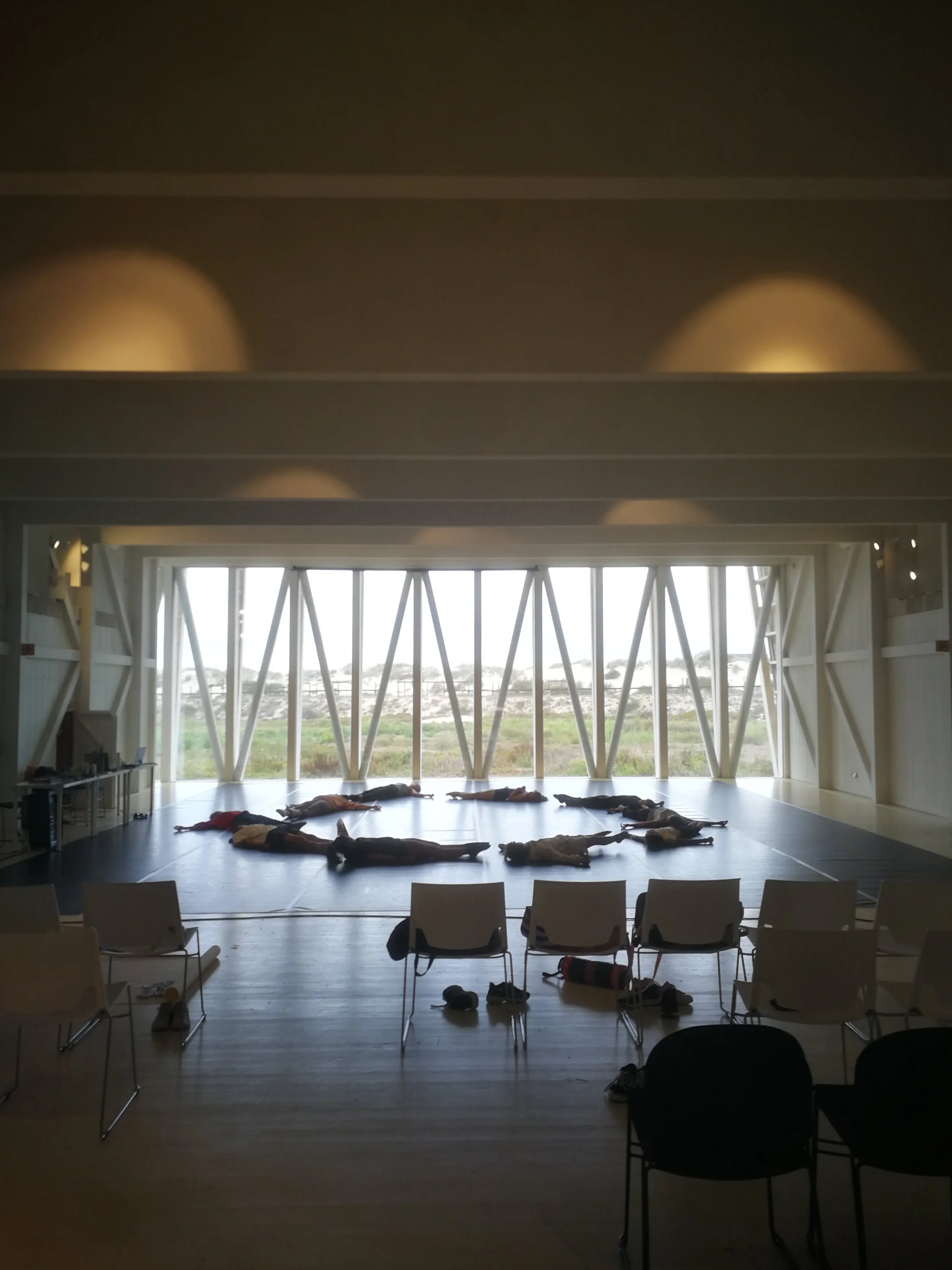 People lying on the floor in a room with large windows revealing an outdoor landscape, chairs arranged around, and some personal belongings scattered on the floor.
