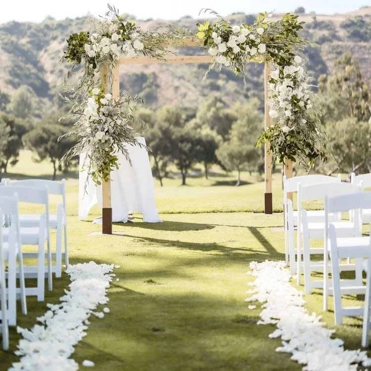 Outdoor wedding ceremony setup with white chairs, a wooden arch decorated with white flowers and greenery, flower petal aisle, and scenic countryside background.