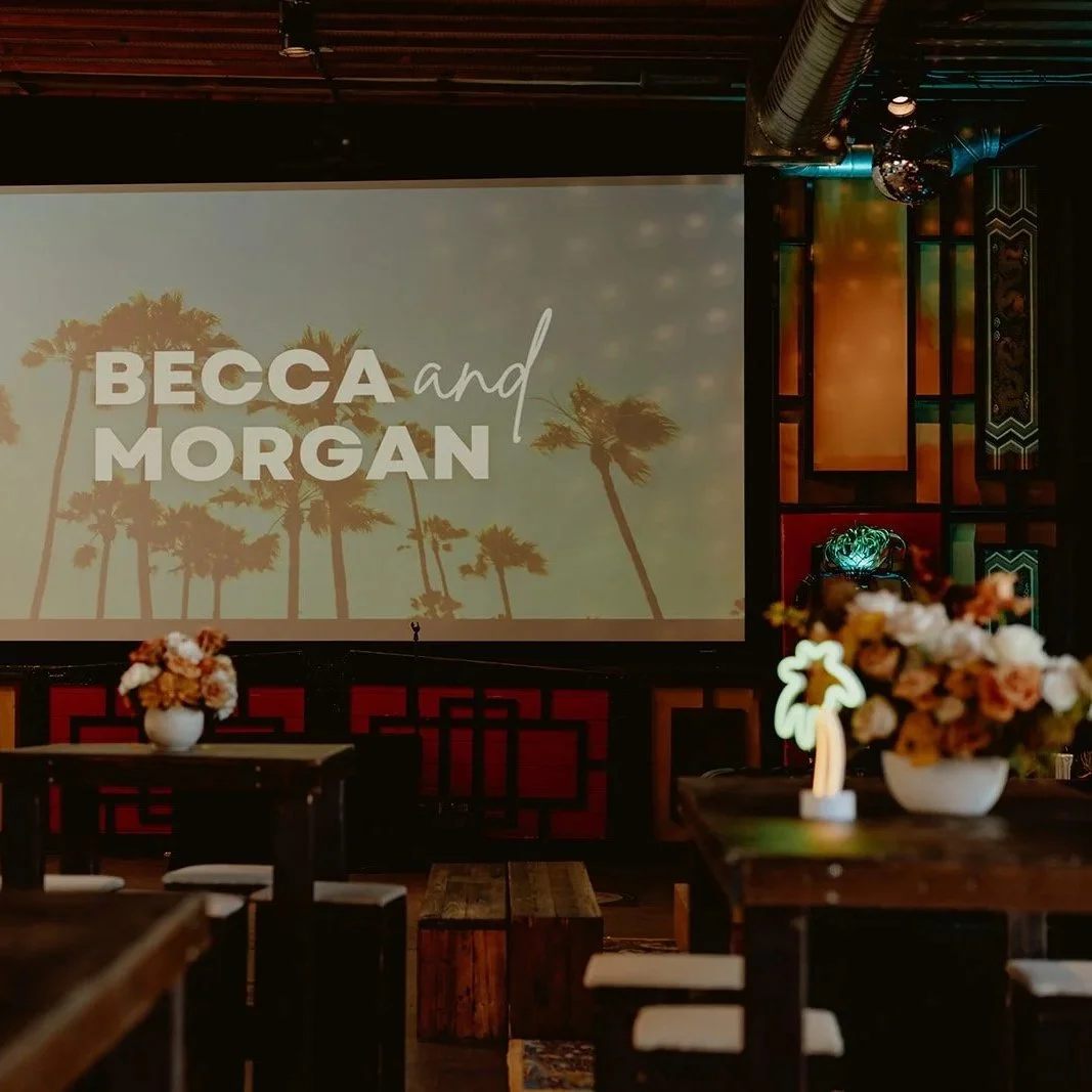 Indoor event space with a large screen displaying 'Becca and Morgan' with silhouettes of palm trees, surrounded by tables with flower arrangements and decorative lighting.