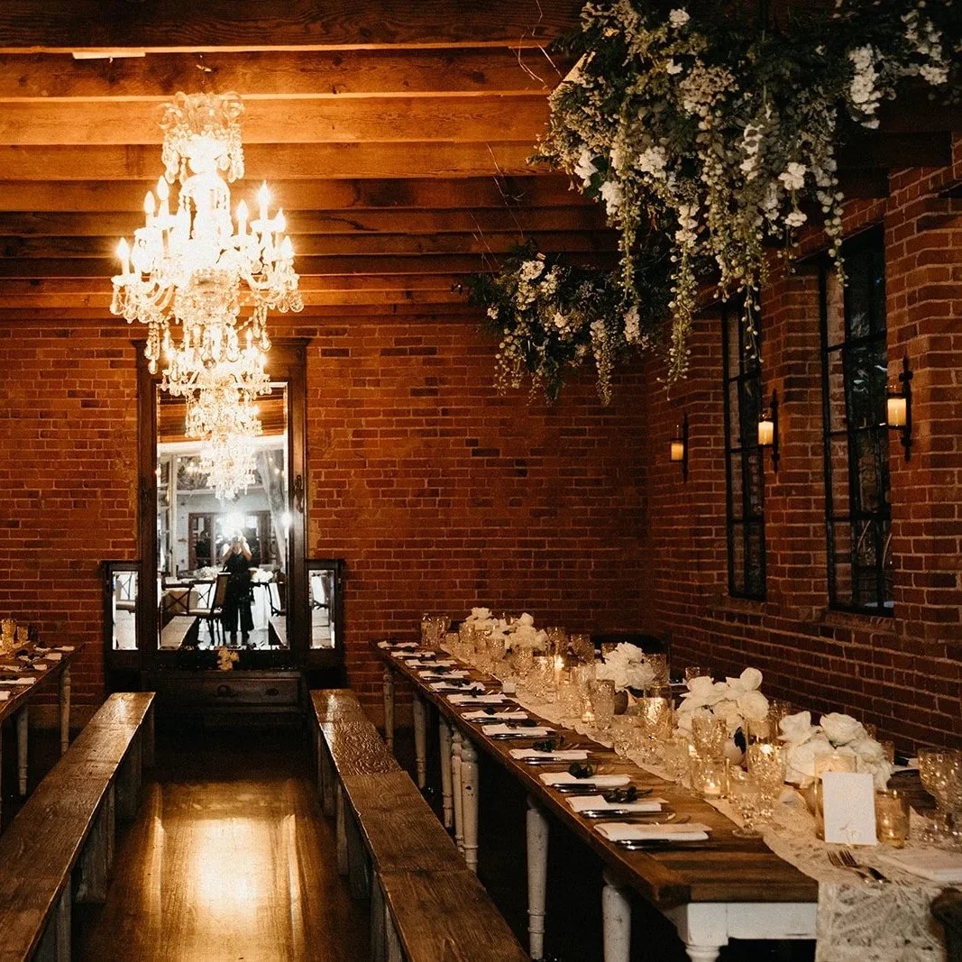 Elegant dining room with brick walls, a large chandelier, floral arrangements, and a long table set for a formal meal.