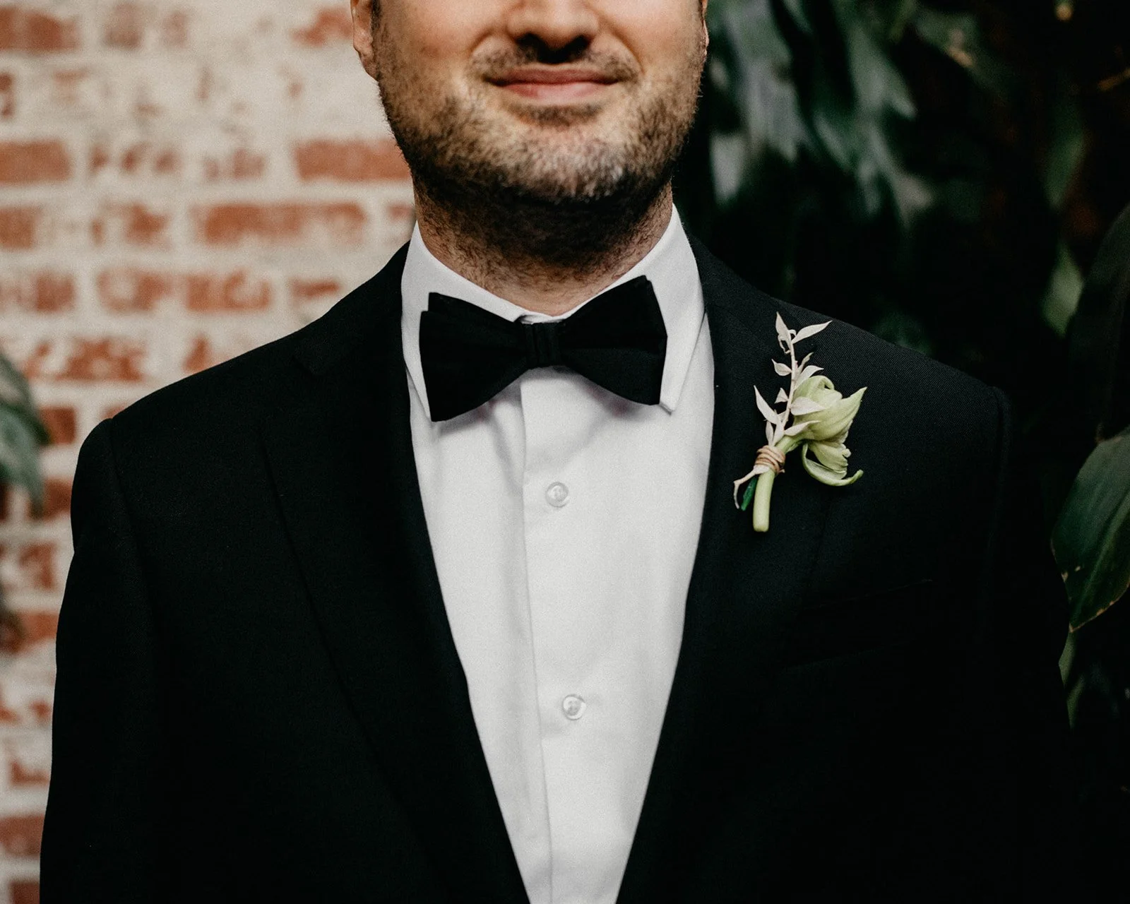Close-up of a man in a tuxedo with a boutonniere, smiling gently, standing in front of a brick wall and greenery.