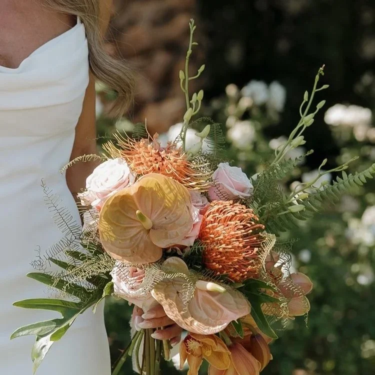 Woman holding a bouquet of peach, pink, and orange flowers with green foliage in an outdoor setting.