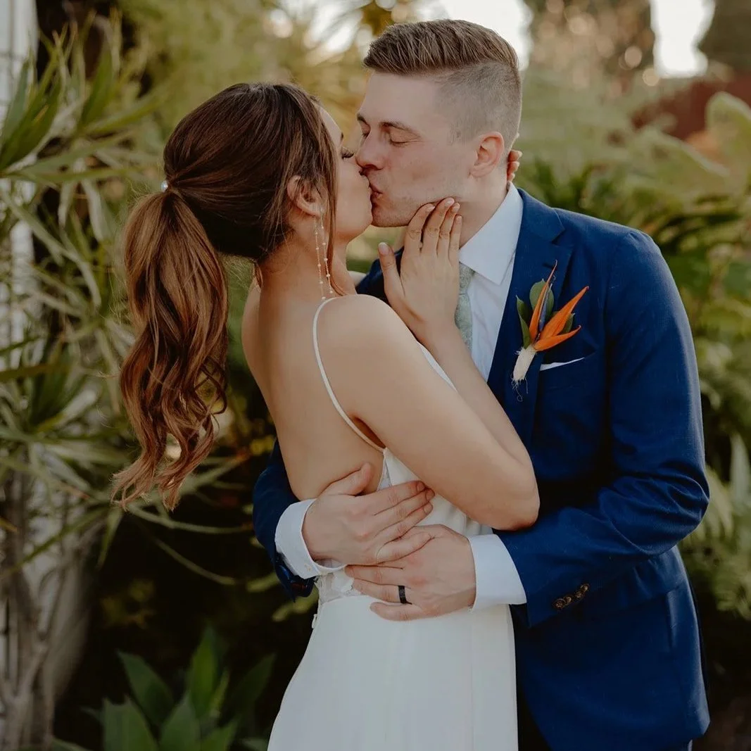 A bride and groom kissing outdoors, surrounded by greenery, during their wedding ceremony.
