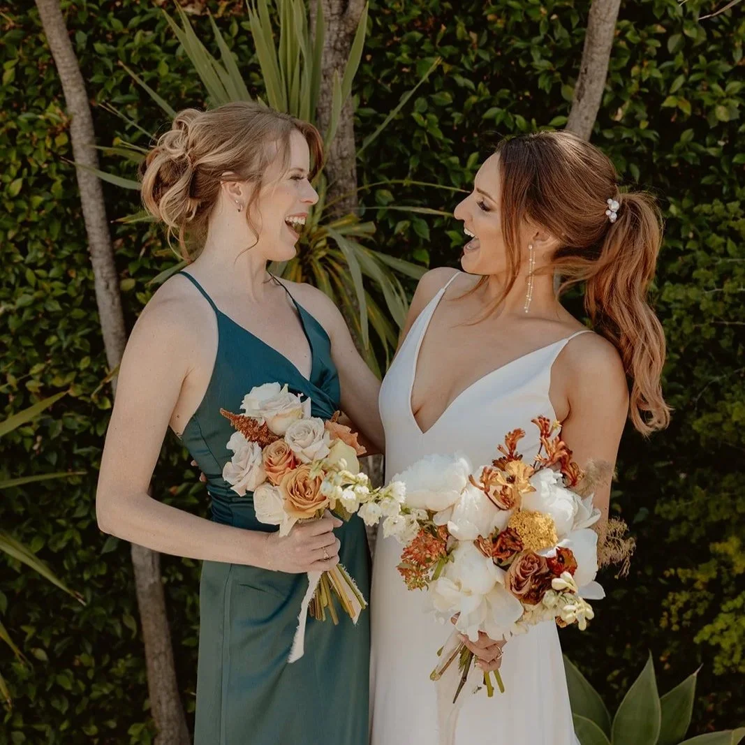 Two women in formal dresses holding bouquets, smiling and sharing a joyful moment outdoors with greenery in the background.