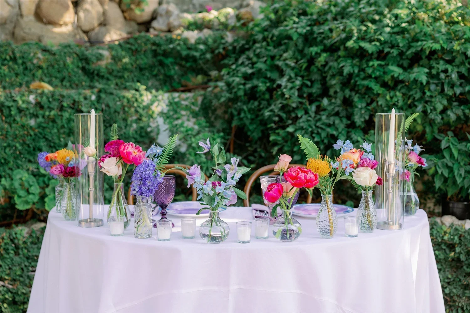 A table set for a celebration or outdoor event, decorated with various colorful flowers in glass vases, surrounded by candles, with greenery in the background.