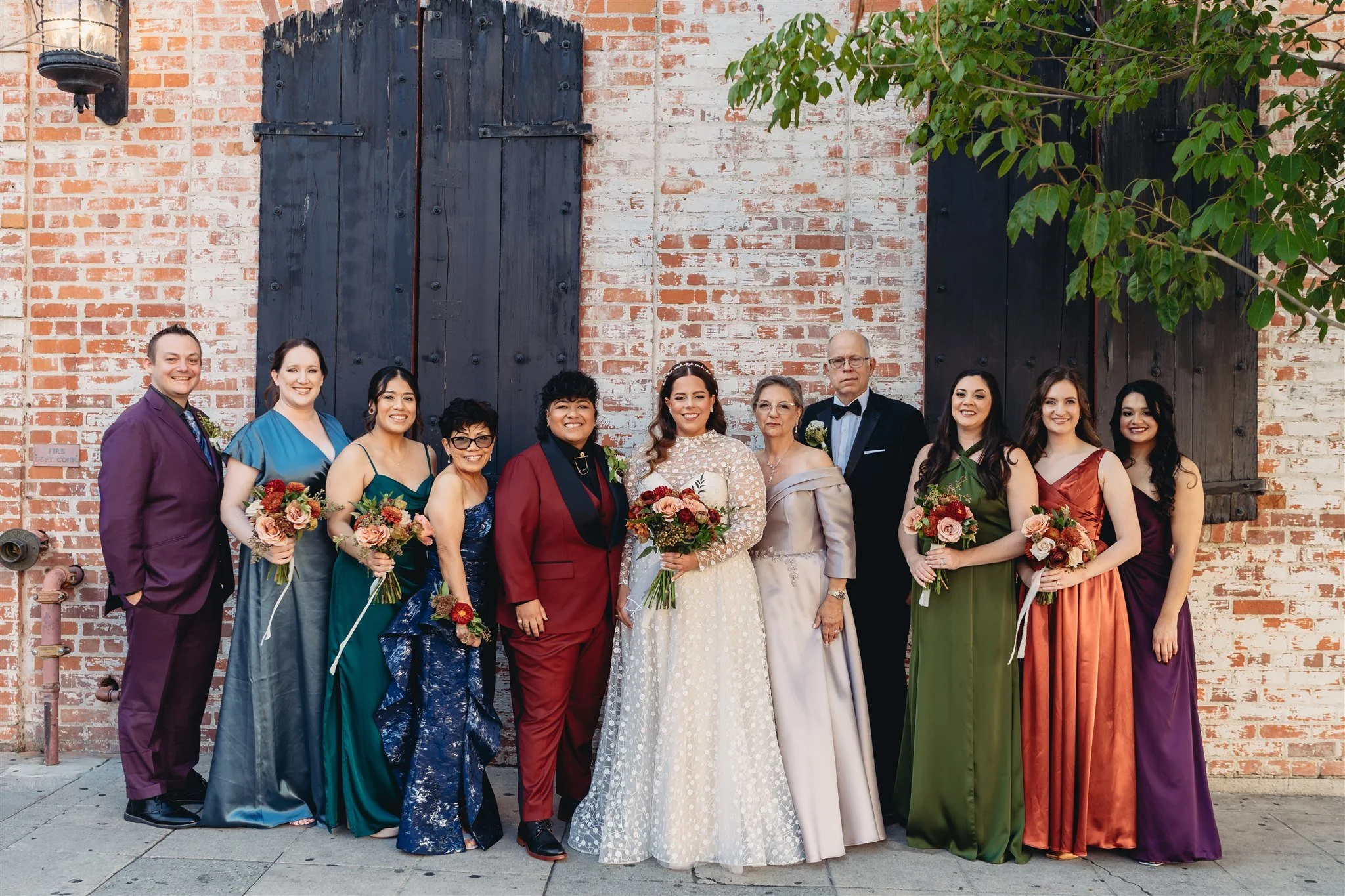 Group of people dressed in formal attire, standing outdoors against a brick wall with black wooden shutters, some holding bouquets of flowers.