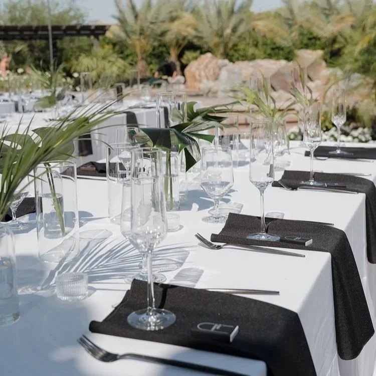 A beautifully set outdoor dining table with glassware, black napkins, and green foliage centerpieces, surrounded by lush garden scenery.