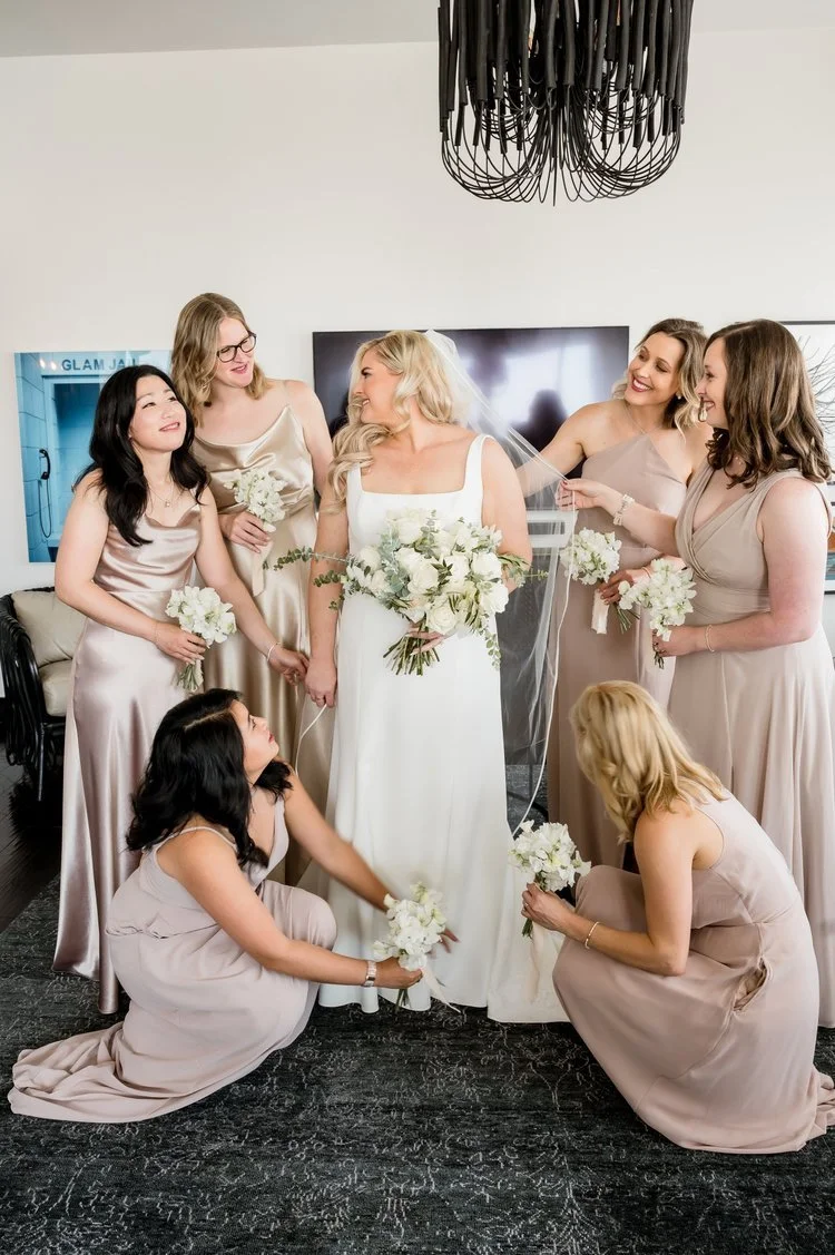 A bride dressed in white with blonde hair holding a bouquet, surrounded by six bridesmaids in pastel dresses, some standing and others crouching, holding white bouquets, all smiling in a well-lit room with a large TV and modern black chandelier overhead.