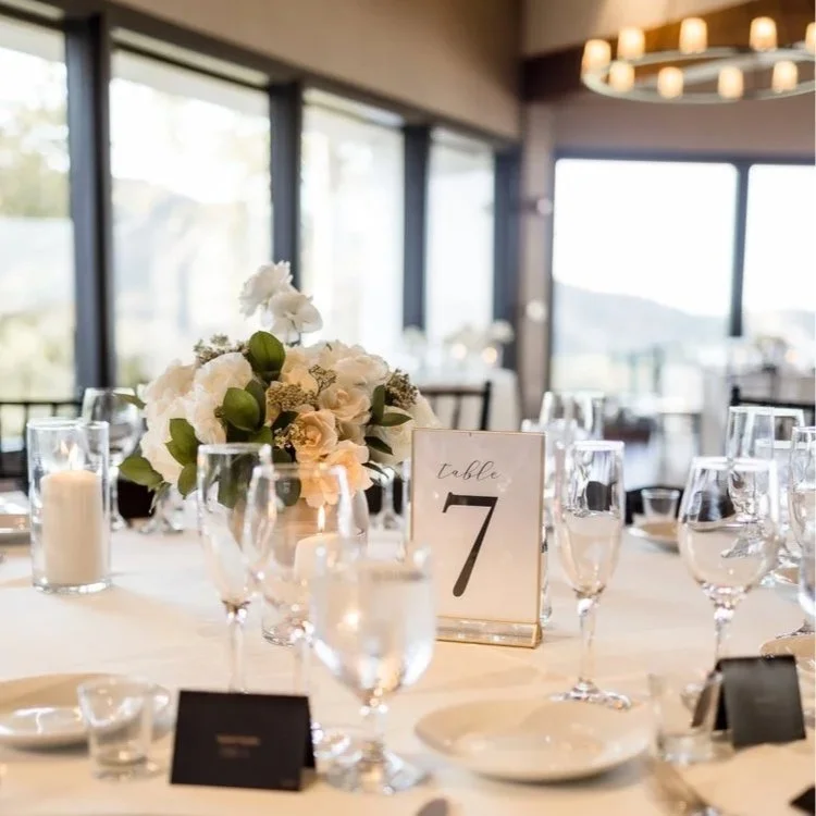 Elegant banquet table set for a wedding or event, featuring a floral centerpiece, table number 7, white plates, glasses, and candles, with large windows and scenic view in the background.