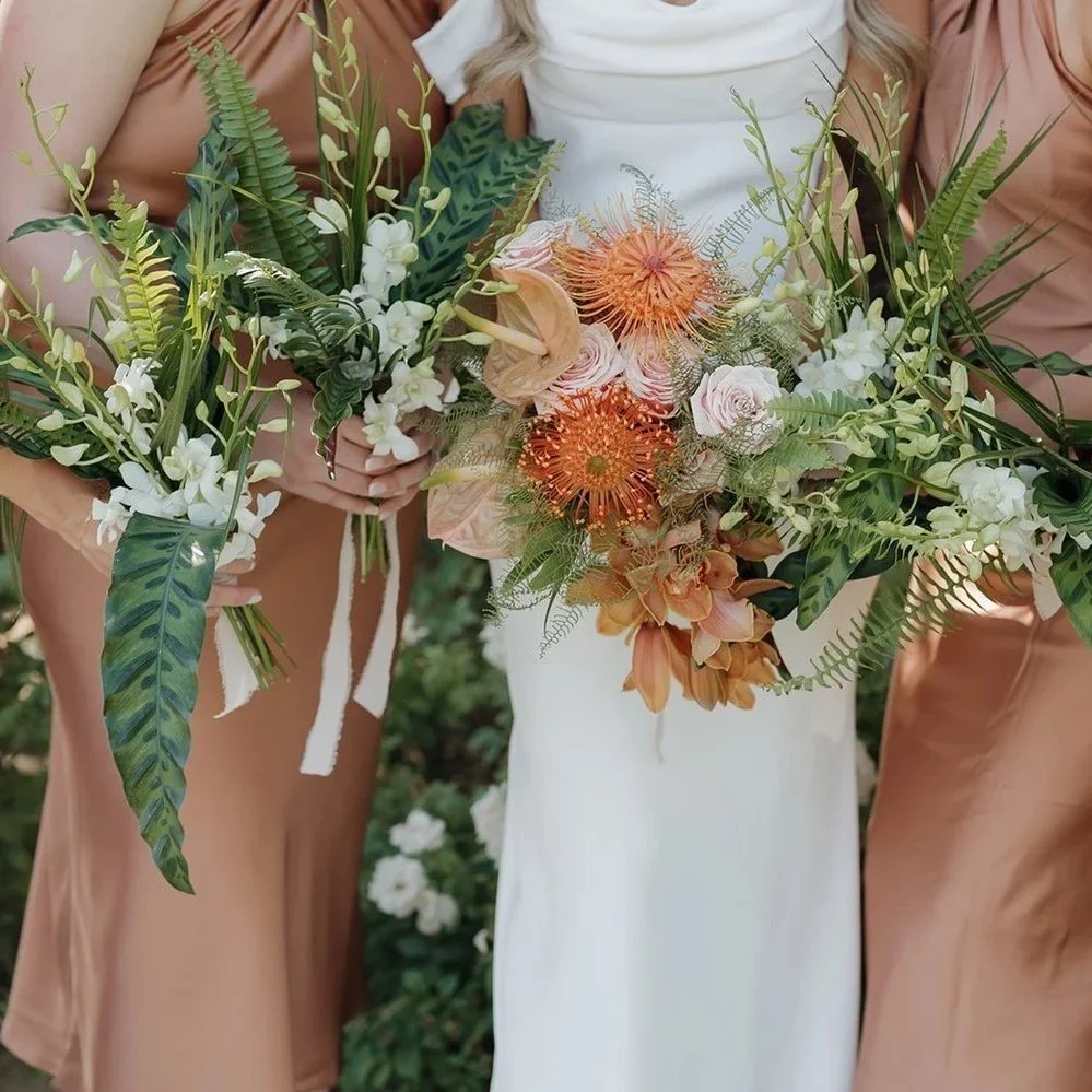 Close-up of three women holding bouquets of flowers, with the middle one in a white dress and the others in bronze-colored dresses, in a garden setting.