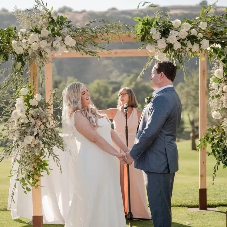 A couple gets married outdoors under a floral arch with a woman officiant in the background, holding hands and smiling at each other.