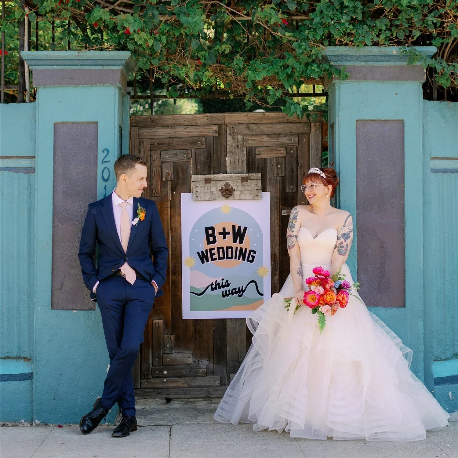 A couple dressed in wedding attire standing in front of a wooden gate with a sign that reads 'B+W Wedding this way'. The groom is in a navy suit with a pink tie, standing with hands in pockets and looking at the bride. The bride is in a strapless white wedding gown with a full skirt, holding a bouquet of colorful flowers, and smiling while looking at the groom.