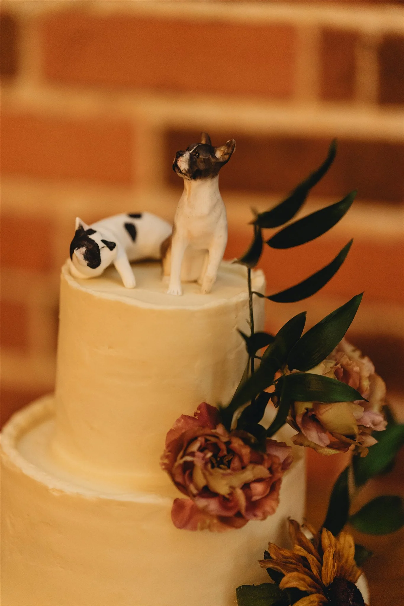 Miniature figurines of two dogs on top of a cylindrical surface, with one standing and the other lying down, surrounded by green leaves and flowers.