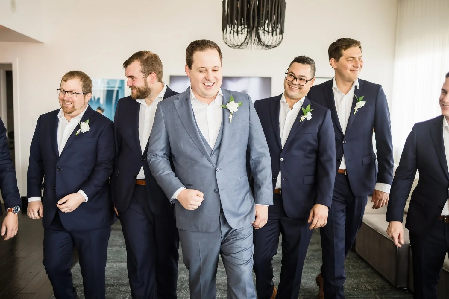Group of men dressed in suits, standing together indoors, smiling, with the central man wearing a light gray suit and the others in dark blue suits with white shirts, some wearing glasses, in a well-lit room with a chandelier and curtains.