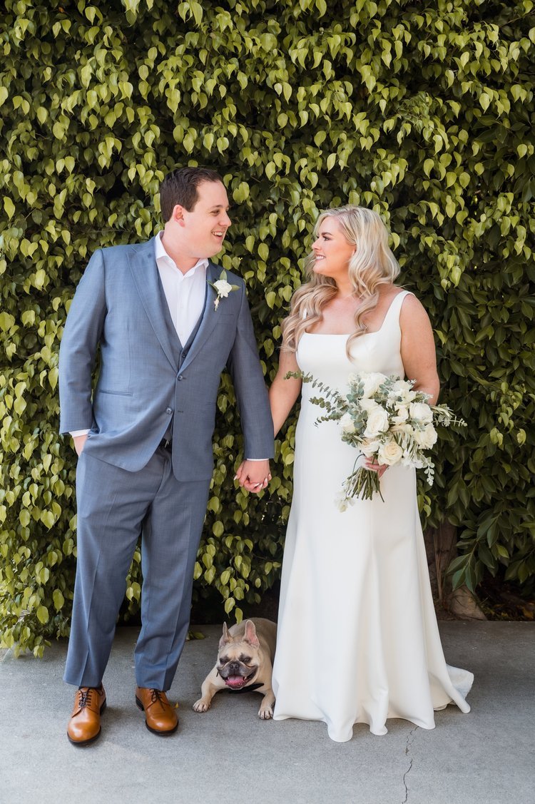A bride and groom holding hands and smiling at each other, standing outdoors in front of a green leafy hedge, with a French bulldog sitting on the ground between them.