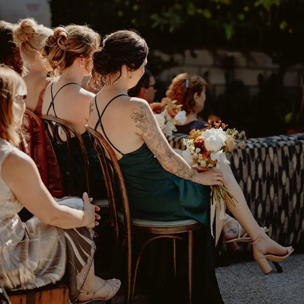 Group of women seated outdoors, some holding bouquets, dressed in formal attire, attending a wedding ceremony.
