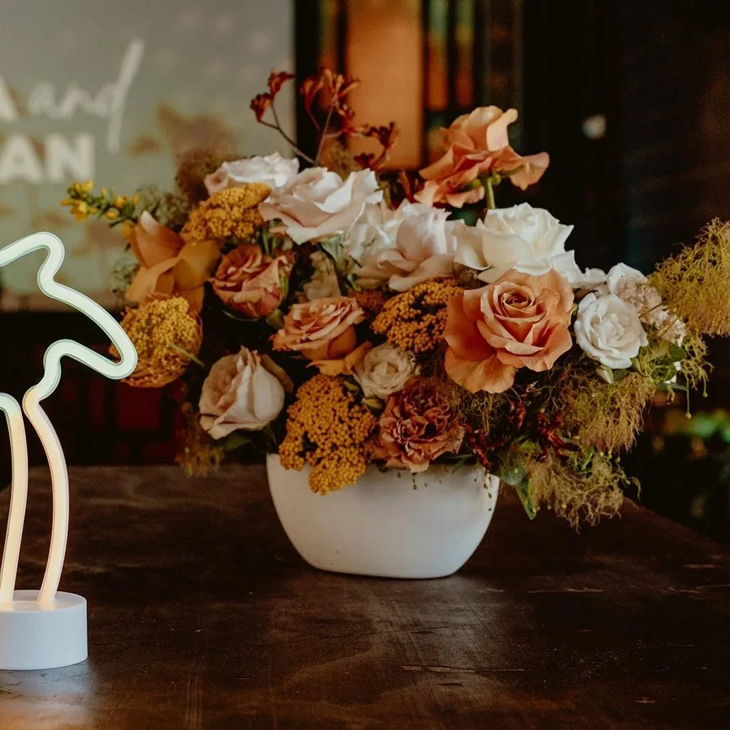 A white vase filled with a mixed bouquet of peach, white, and light pink roses, yellow and orange filler flowers, and green foliage on a dark wooden table.