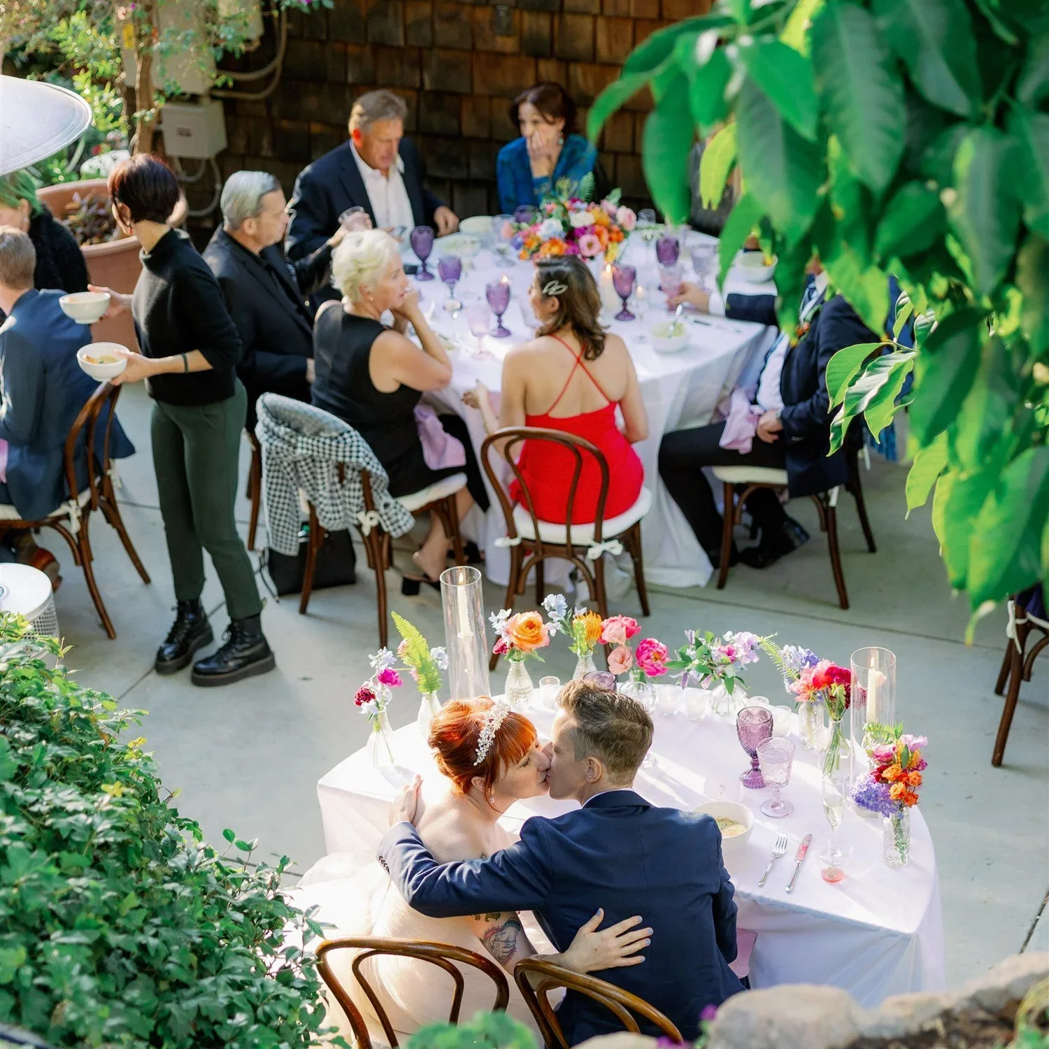 A wedding reception outdoors with guests sitting at decorated tables, including a couple kissing in the foreground, and a group seated at a round table in the background, surrounded by greenery and flowers.